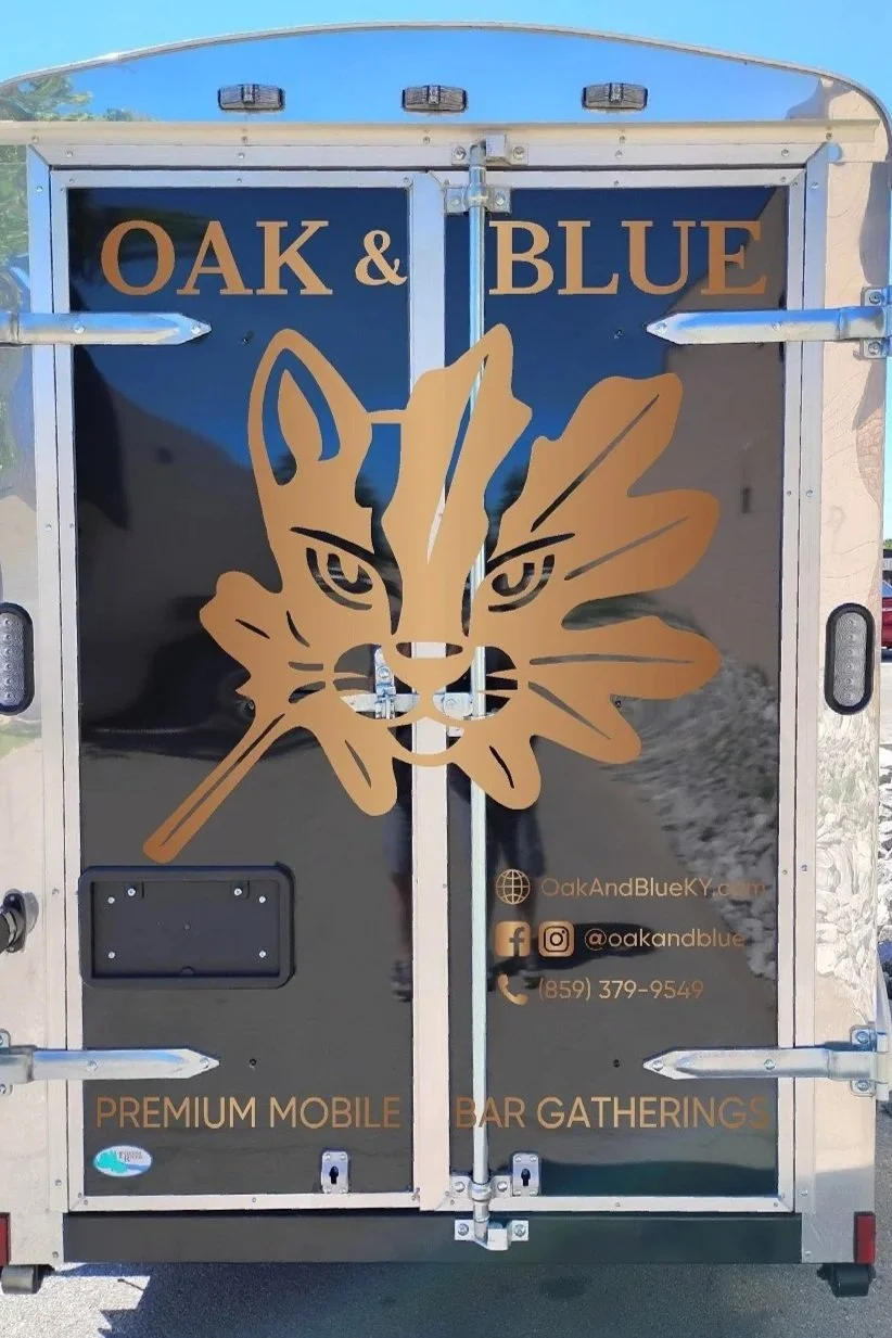 The back of a mobile bar trailer with the name "Oak & Blue" and a graphic of a feline face with a leaf behind it. Contact information and social media handles are also displayed, with the phrases "Premium Mobile" and "Bar Gatherings".