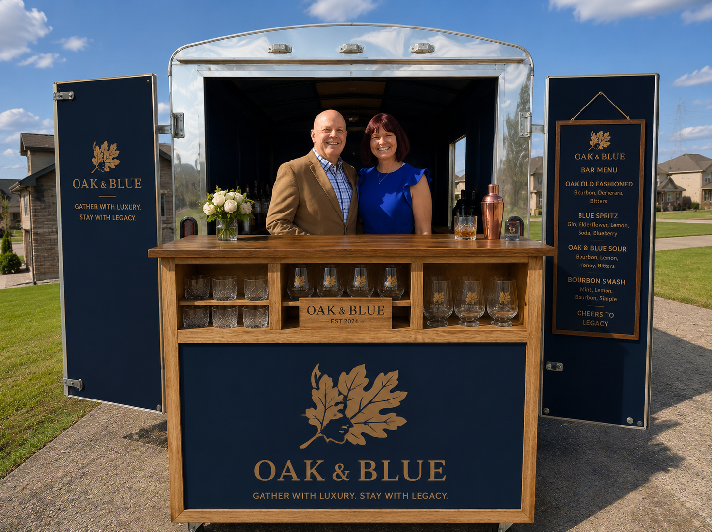 A man and woman standing behind a mobile bar called OAK & BLUE, outdoors in a neighborhood yard under a blue sky with a few clouds. The bar has a wooden countertop, glasses with a leaf logo, and a sign with the bar menu. The man is wearing a brown blazer and checkered shirt, and the woman is wearing a blue dress.