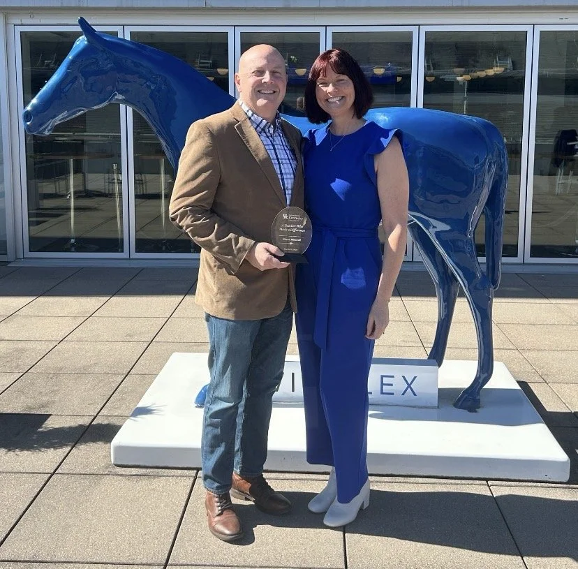 A man and woman standing side by side outside in front of a glass building, smiling at the camera. The man is holding an award plaque. Behind them is a large blue horse sculpture. The woman is dressed in a blue jumpsuit, and the man is wearing a brown blazer, checkered shirt, and jeans.