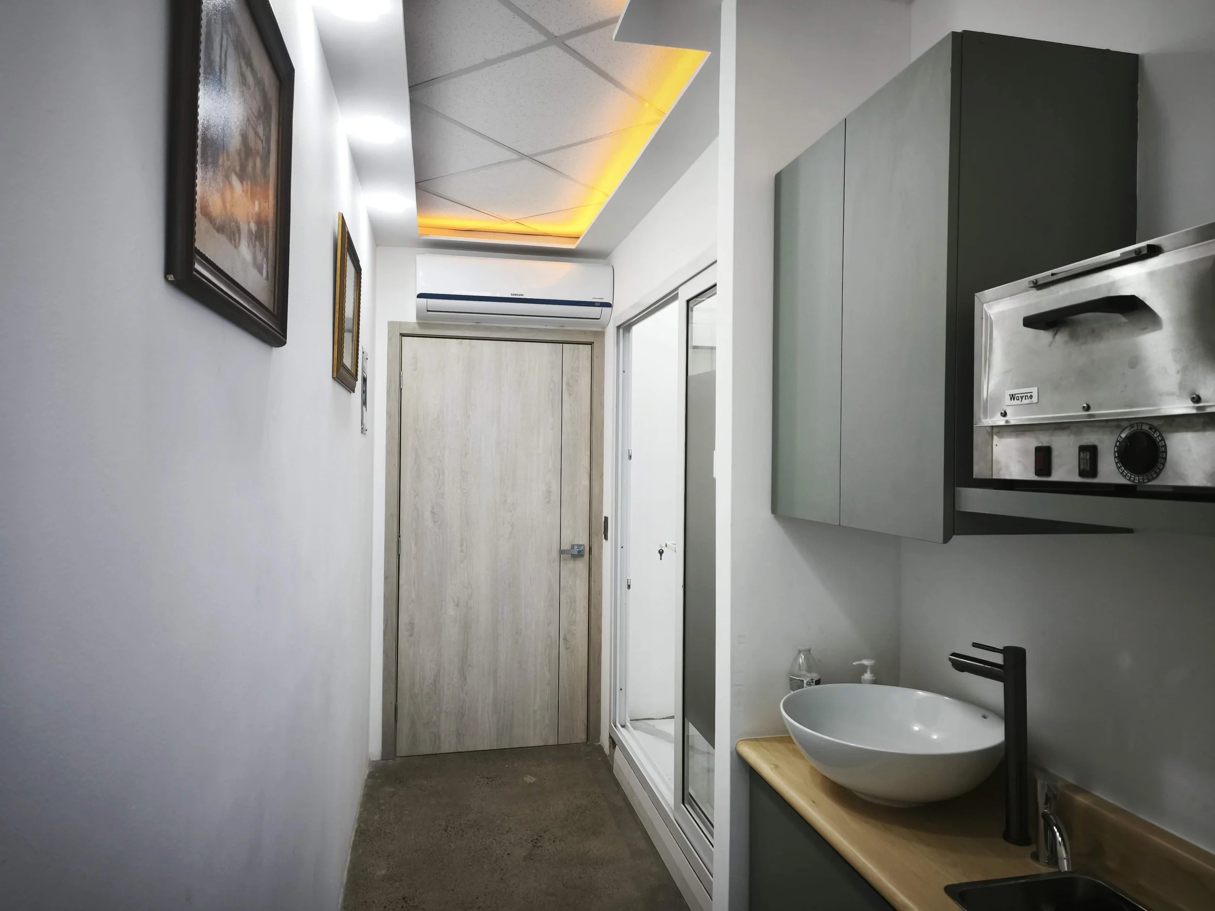 Interior de una pequeña oficina o cuarto, con pared blanca, una puerta de madera en la parte trasera, aire acondicionado blanco en la parte superior de la puerta, un armario gris en la parte derecha, una ventana al lado del armario, lavabo con grifo negro, y en la esquina inferior derecha un dispensador de jabón y un envase de agua.