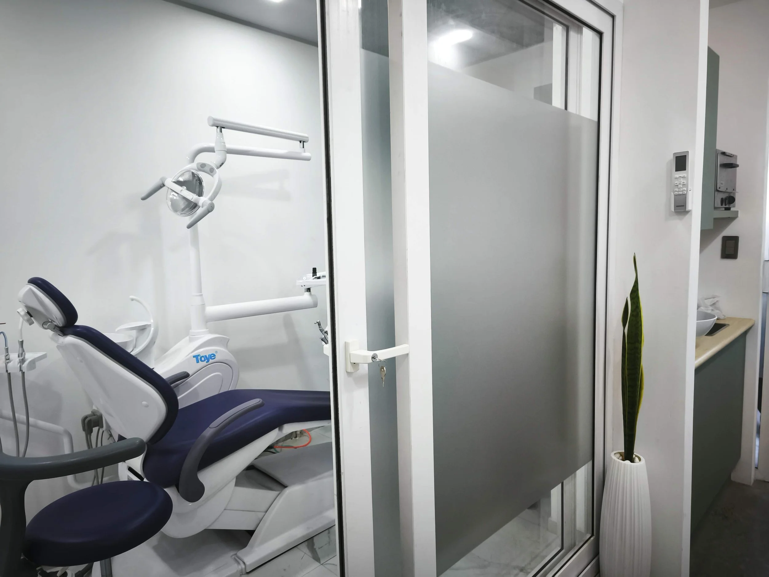 Sala de consulta en clínica dental con sillón dental, lámpara y equipo en la esquina, vista a través de una puerta de cristal.