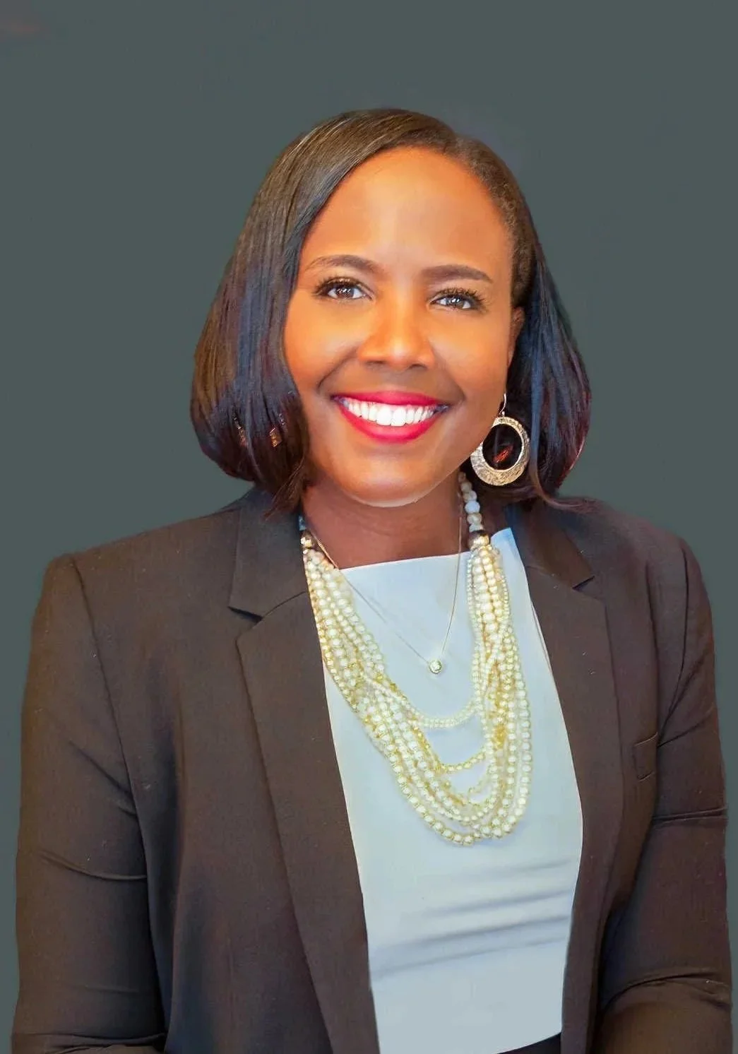 A woman with shoulder-length black hair, wearing a black blazer, white top, and layered pearl necklaces, smiling against a gray background.