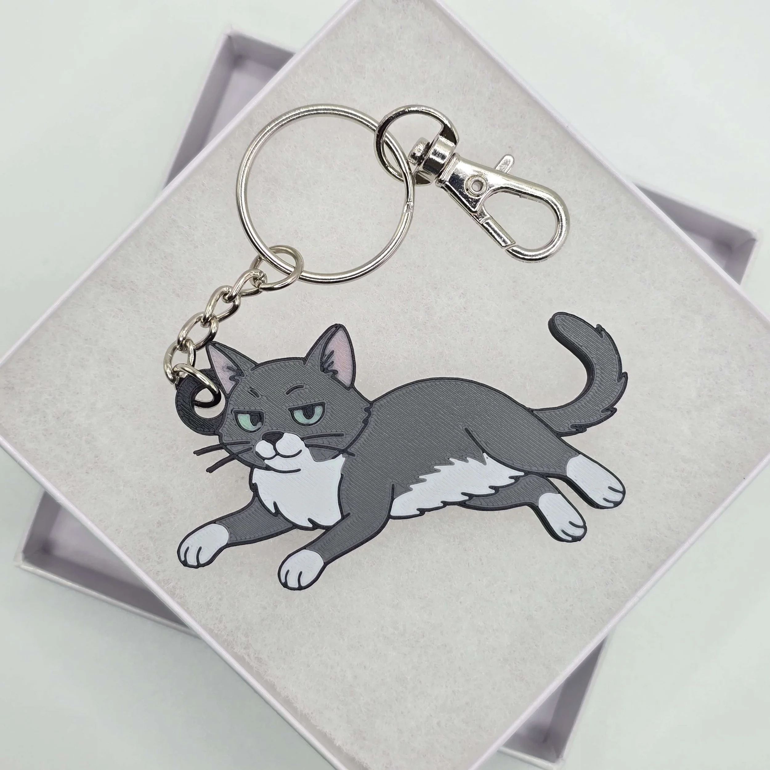 A silver keychain with a carabiner clip attached to a circular ring, resting on a white surface with a cartoon gray cat sticker nearby.