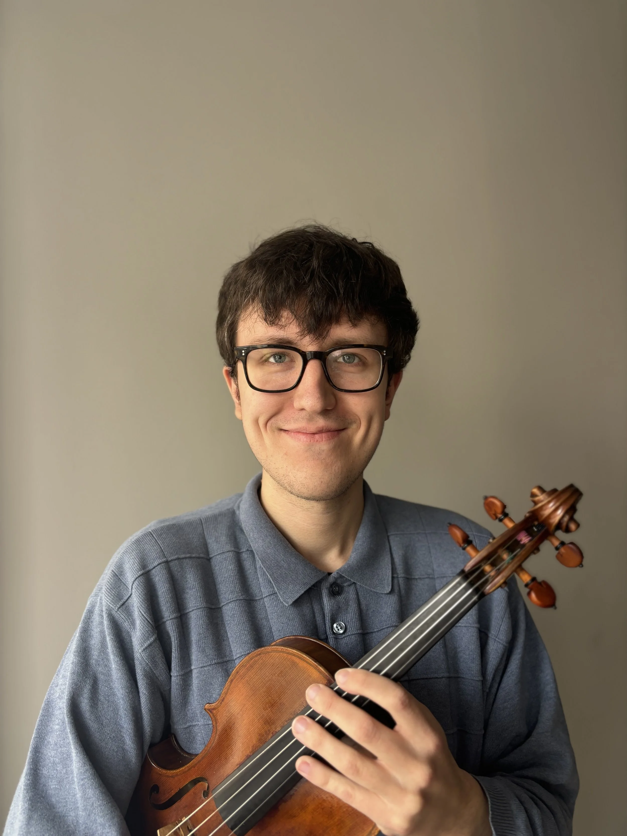 A young man with glasses, wearing a gray collared shirt, is smiling while holding a violin.