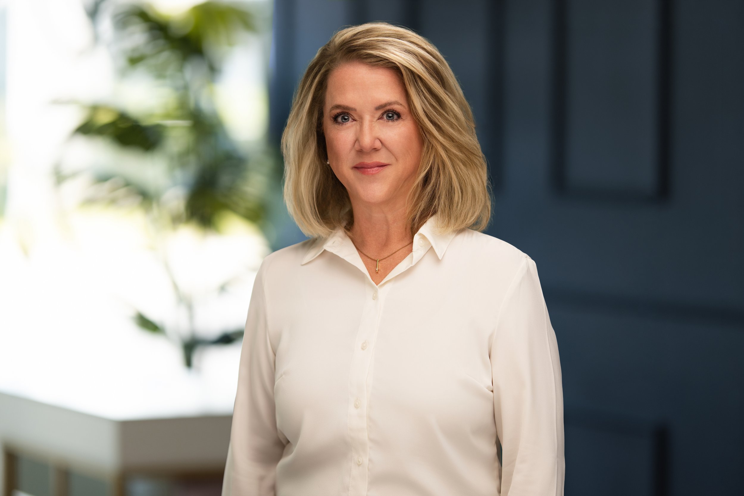 A middle-aged woman with shoulder-length blonde hair, wearing a white button-down shirt, standing in a modern office environment with blurred plant in the background.