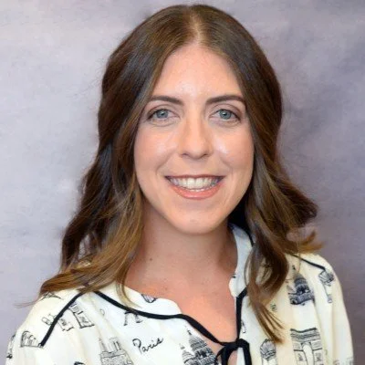 A woman with long brown hair, blue eyes, and a bright smile, wearing a white shirt with black and gray sketches and a black trim, standing against a gray background.