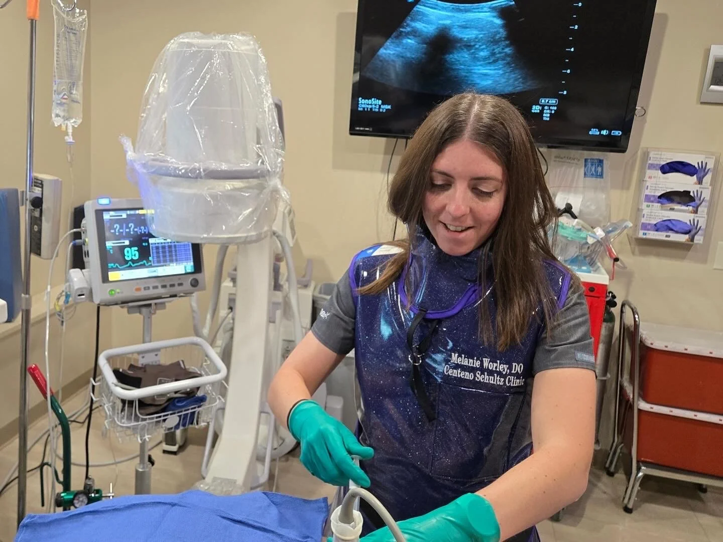 A woman medical professional in scrubs and green gloves working in a hospital room with medical equipment, monitors, and an ultrasound screen displaying a scan.