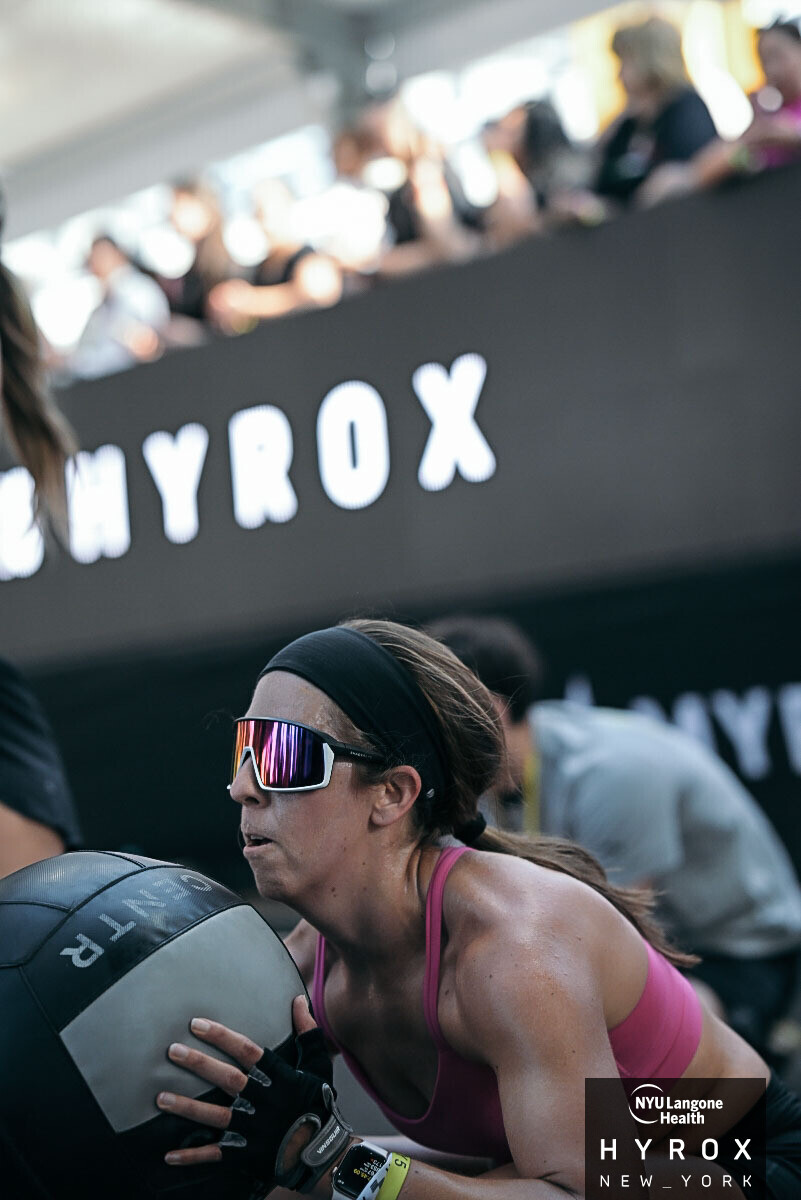 Woman wearing sports goggles and a pink sports bra holding a medicine ball during a workout in a gym with a HYROX banner in the background.