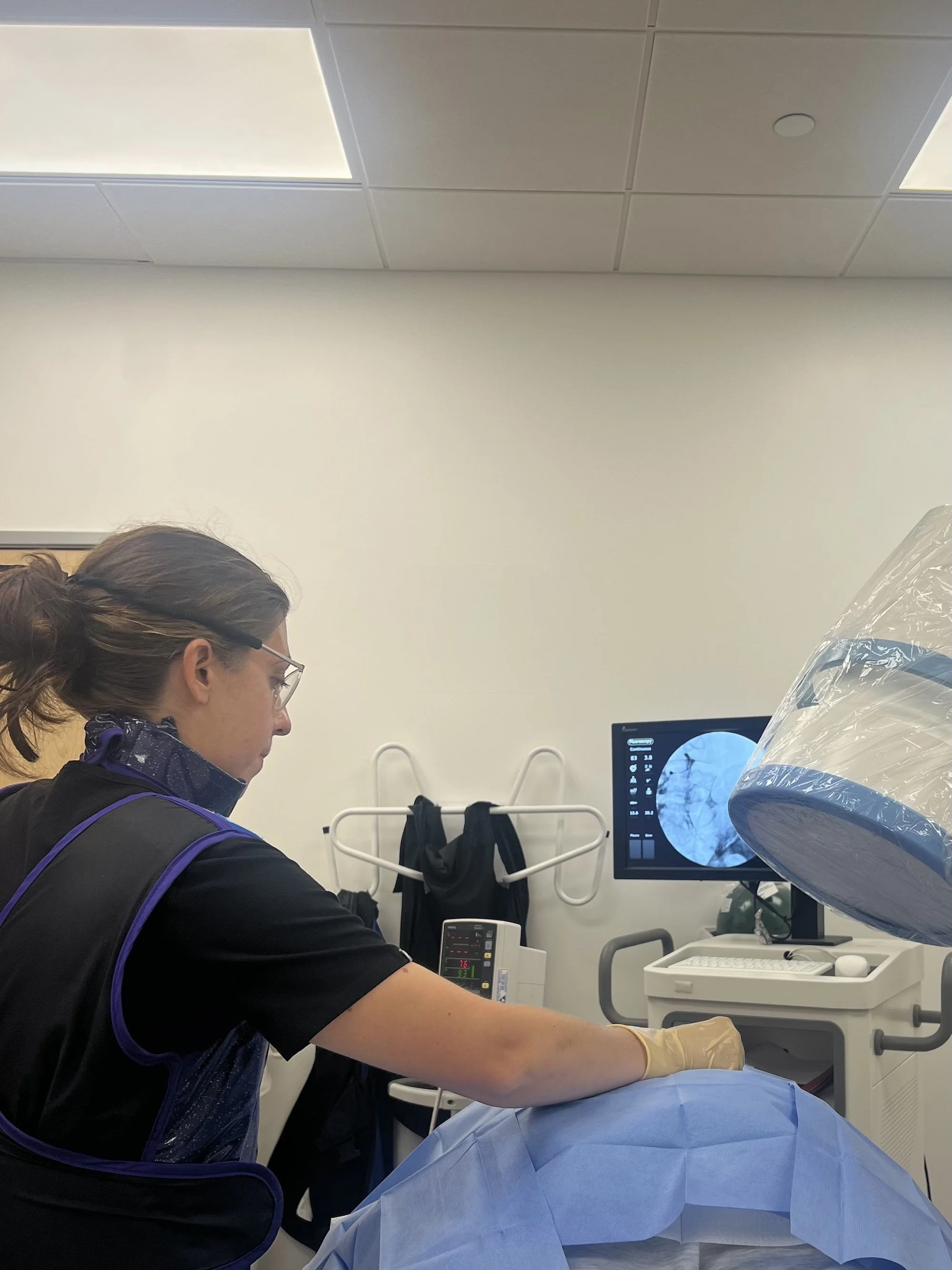 Medical professional in scrubs and gloves performing a procedure on a patient in a hospital or clinic room, with a monitor displaying a medical image.