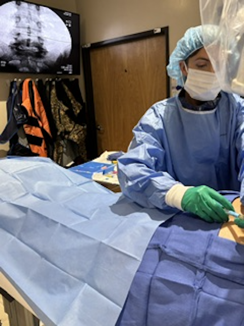 A healthcare worker in surgical attire, including a mask, gloves, gown, and face shield, is performing a medical procedure in a hospital or clinic room. An X-ray image is displayed on a monitor in the background.