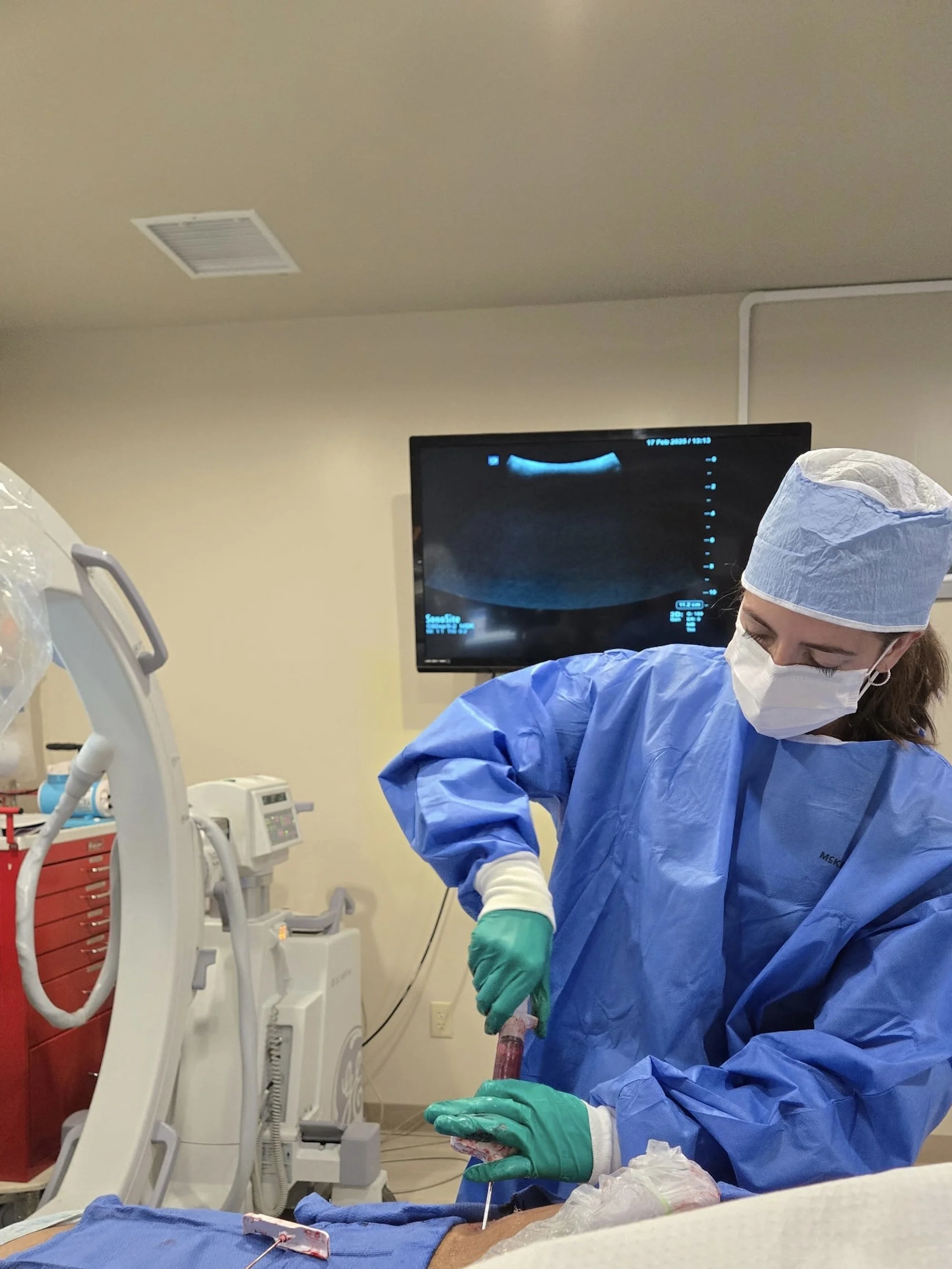 Medical professional in blue scrubs, face mask, and green gloves performing a procedure in a hospital room with medical equipment and monitor in the background.