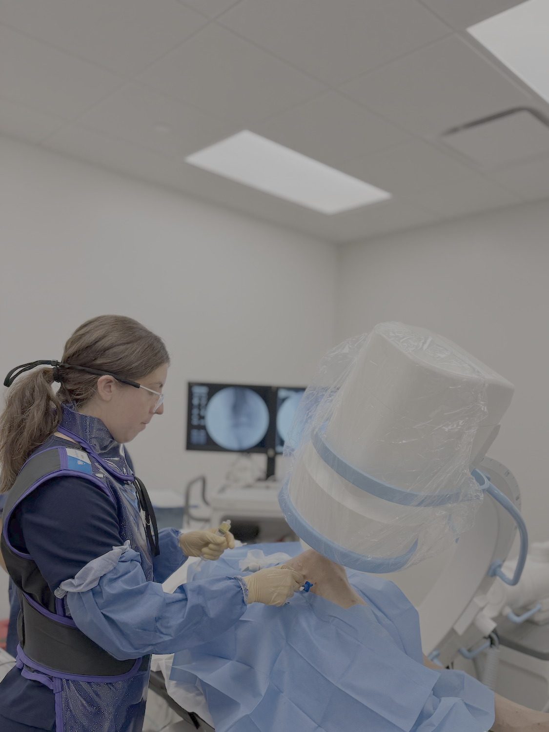 A female physician in scrubs and gloves assisting a patient in a hospital room with medical equipment and monitors behind her.