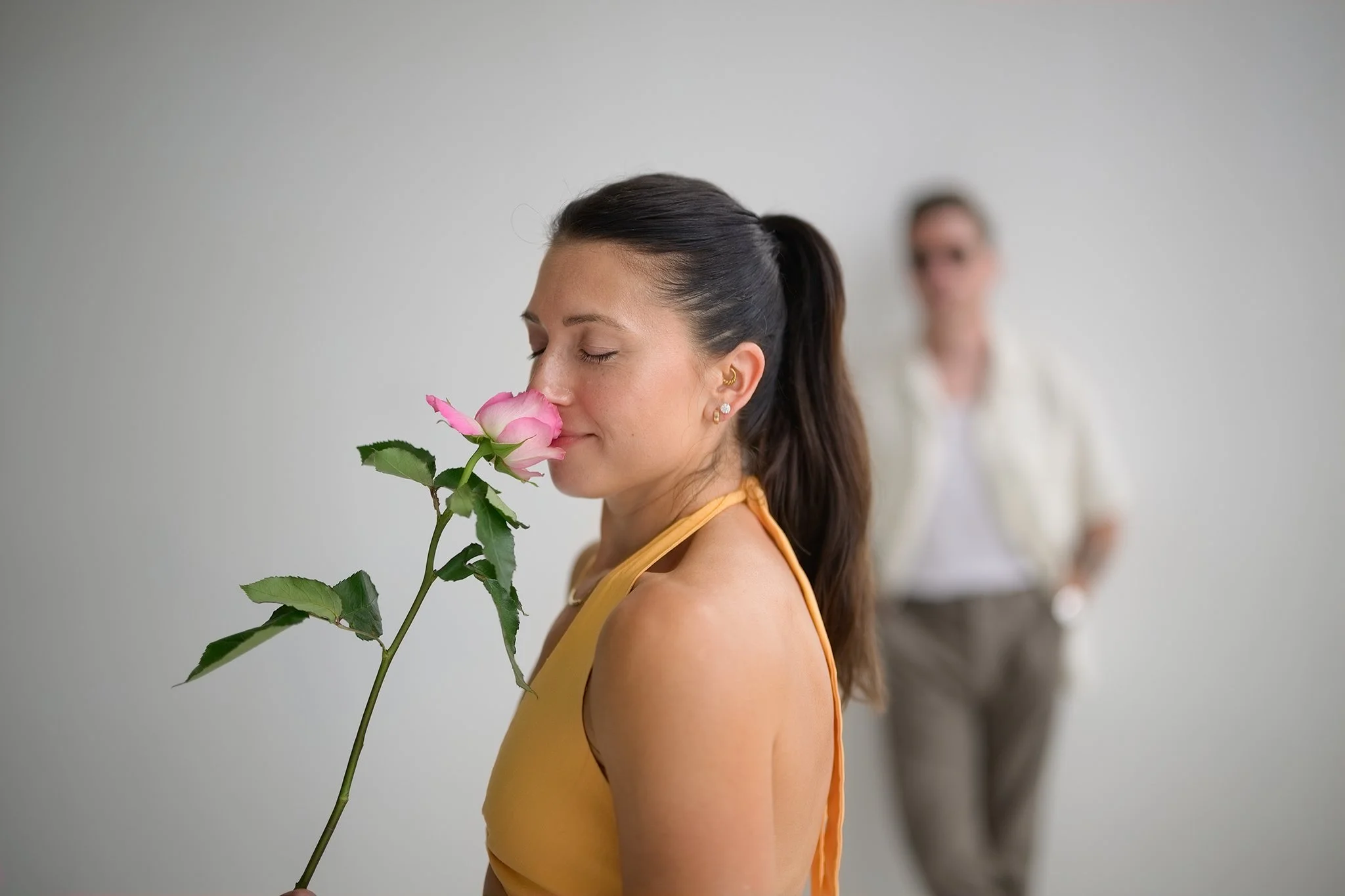 A woman with long dark hair, wearing a yellow top, smelling a pink rose on a stem with green leaves, with a blurred woman in the background standing against a plain white wall.