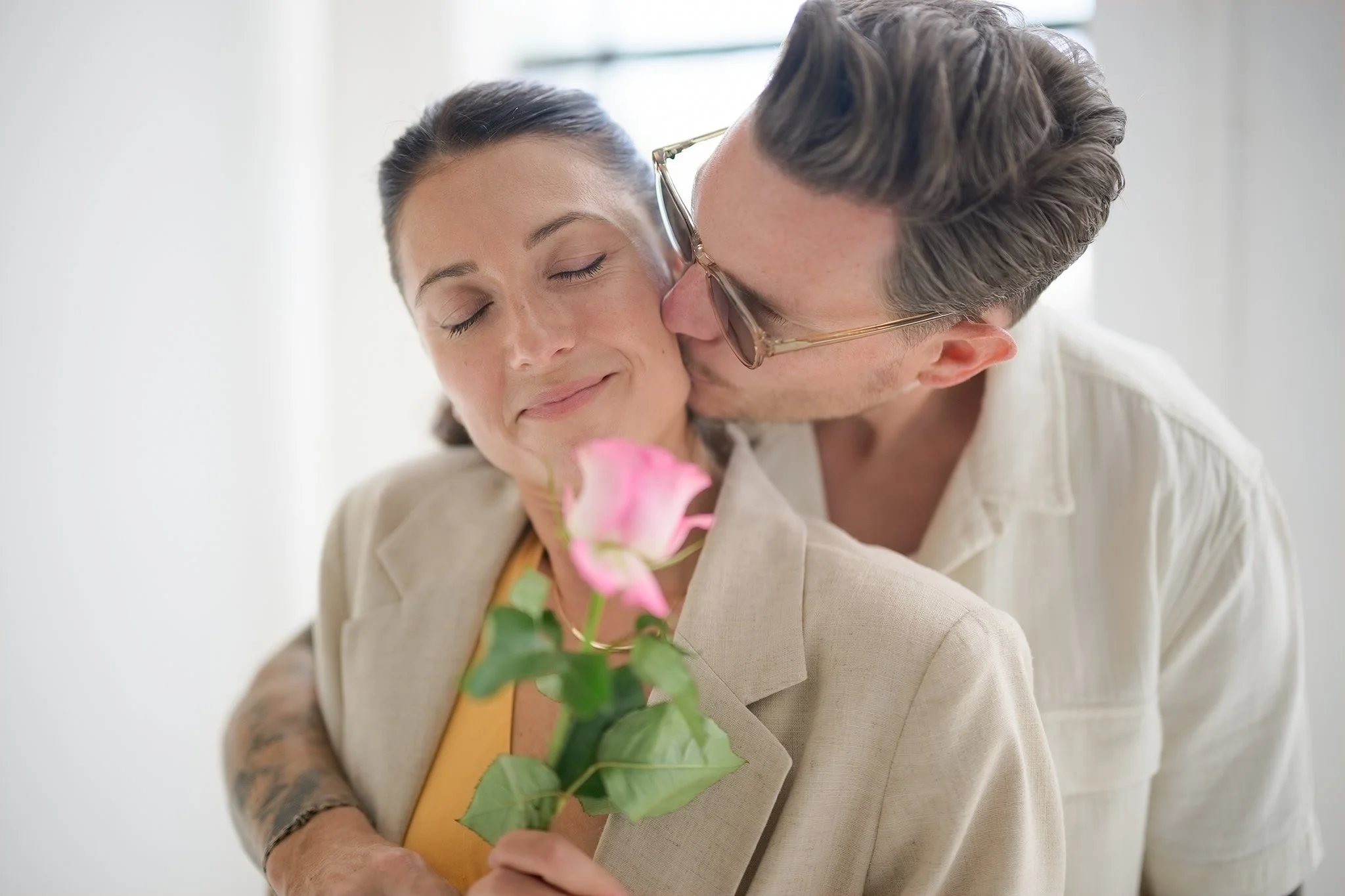 A man wearing glasses and a cream suit is giving a kiss on the cheek to a woman with her eyes closed, holding a pink flower with green leaves.