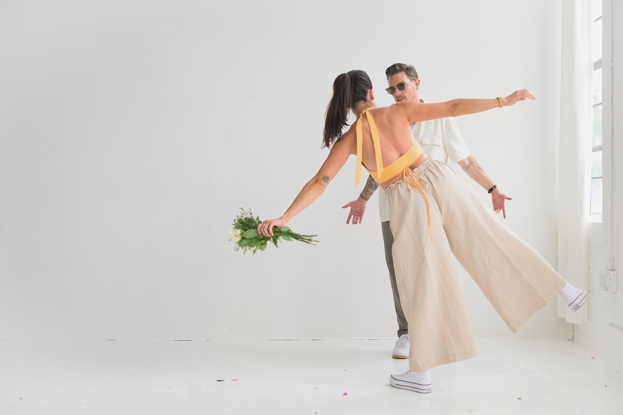 A woman in yellow top and beige wide-leg pants dancing with a man in sunglasses inside a bright white room, holding a bouquet of flowers in her left hand.