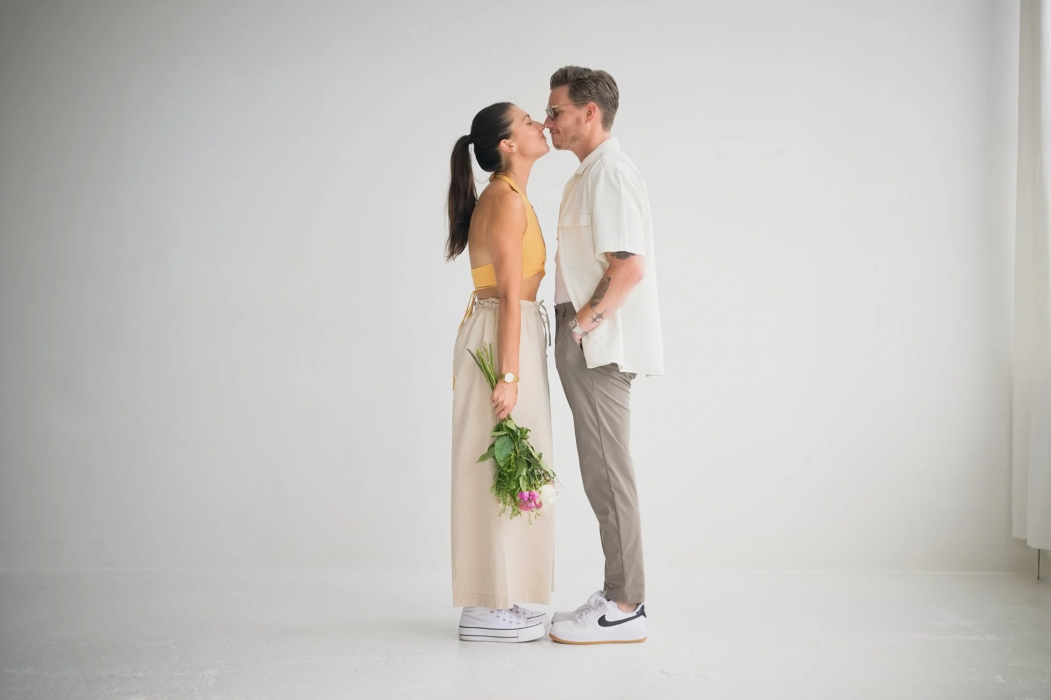 A couple standing close face-to-face with noses touching, holding a bouquet of pink flowers with green leaves, in a minimal white space. The woman is wearing a yellow crop top and beige pants, holding the bouquet in her left hand. The man is wearing 