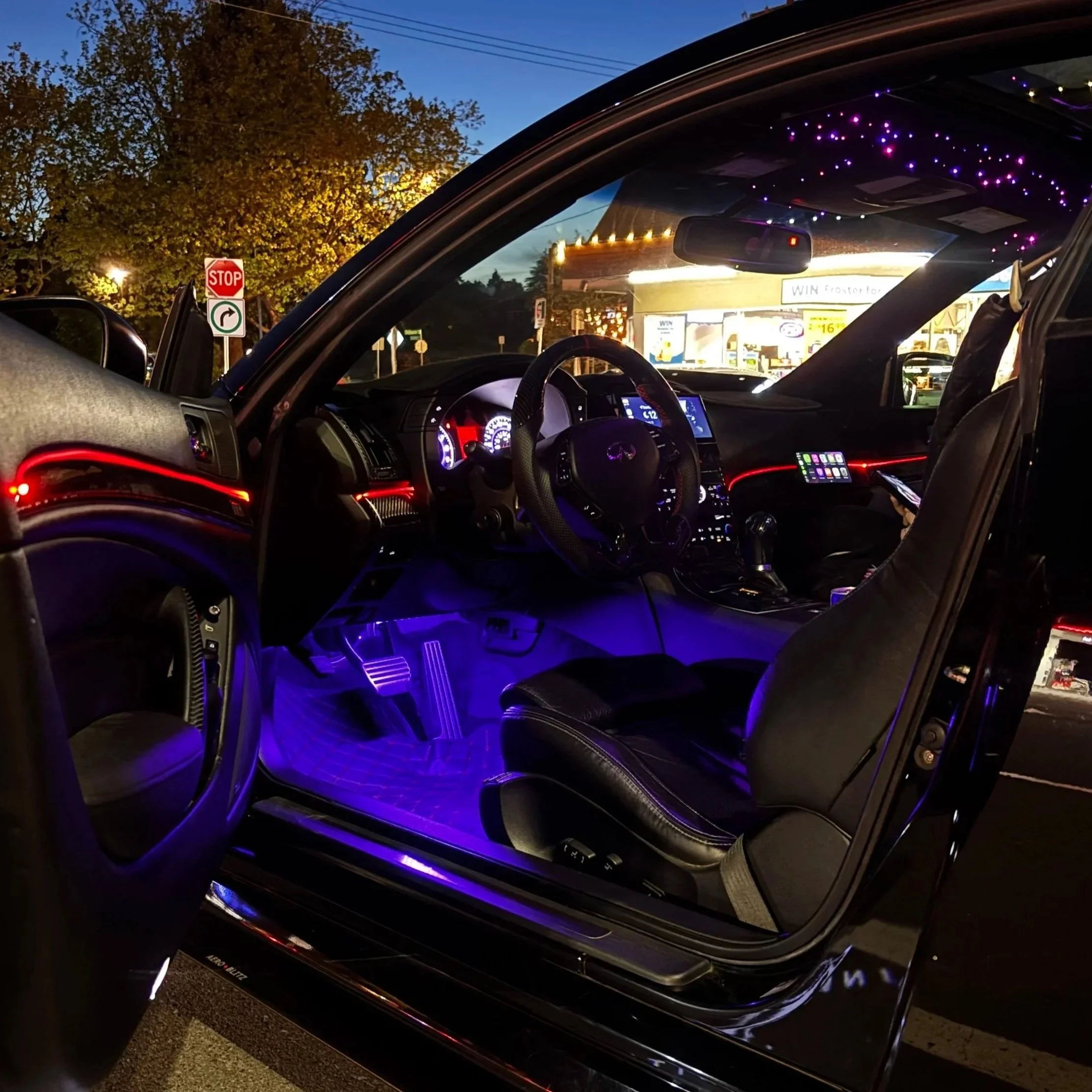 The photograph captures the interior of a car at sunset, showing illuminated dashboard instruments, purple and red ambient lighting, and the view through the windshield of a stop sign, a right turn sign, power lines, a tree with leaves, and a commercial building with bright signage.
