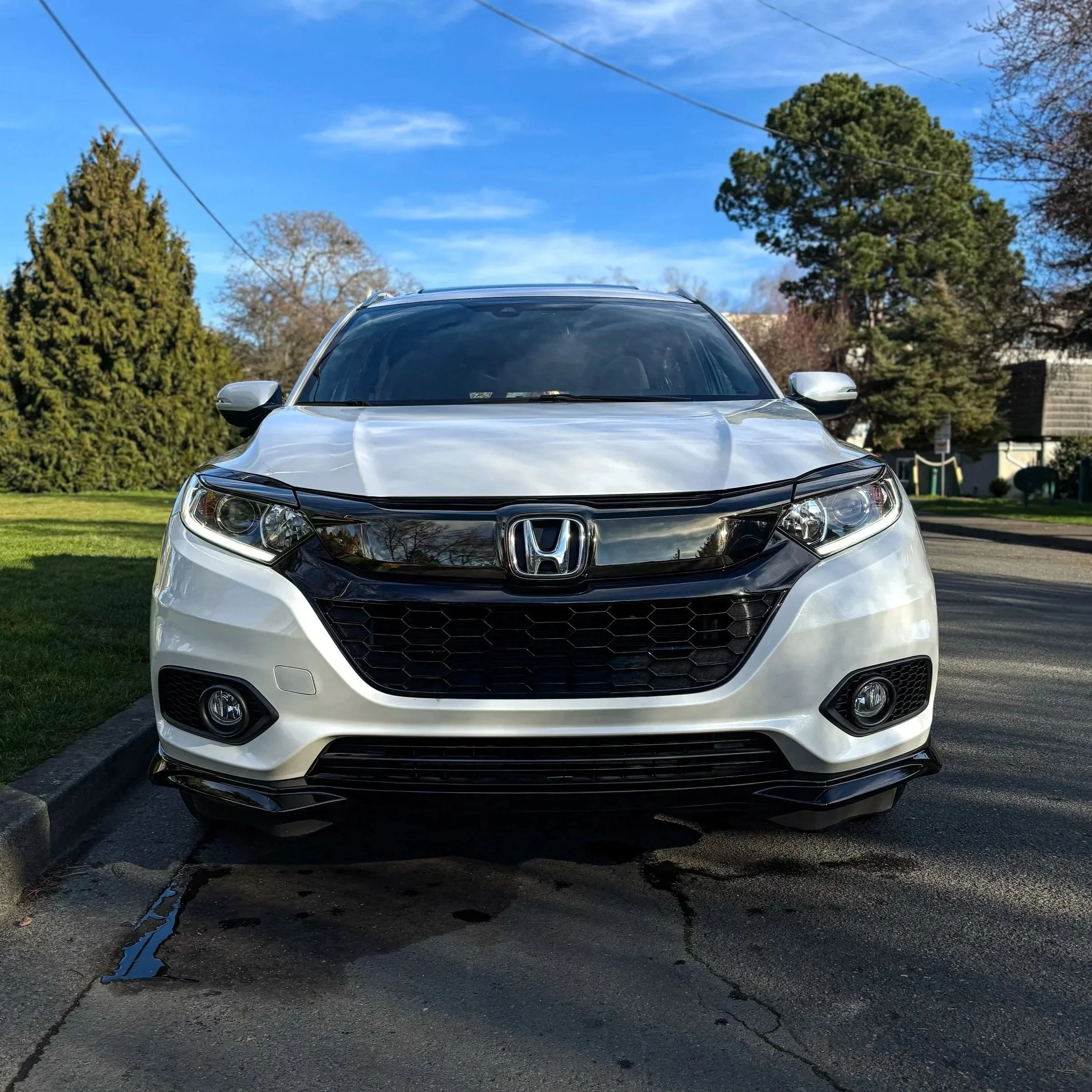 A silver Honda car parked on a residential street with trees and houses in the background under a blue sky.