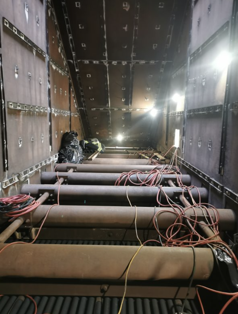 View inside a large industrial or construction vehicle with black metal walls, pipes, and tangled wires, with bright lights illuminating the interior.
