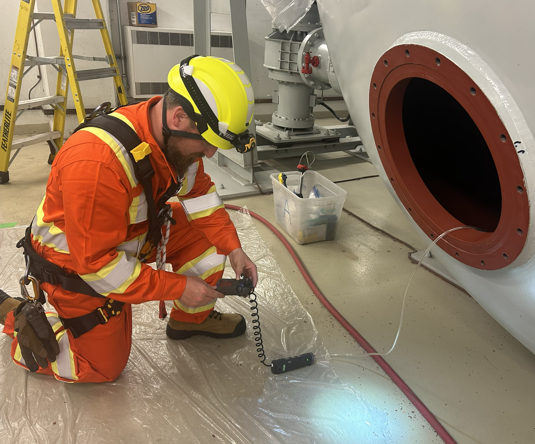 A worker in orange safety gear and yellow helmet kneeling on the floor, working near a large piece of industrial equipment. The worker is holding an air monitor sampling air within a confined space prior to a worker entering to ensure the air is safe
