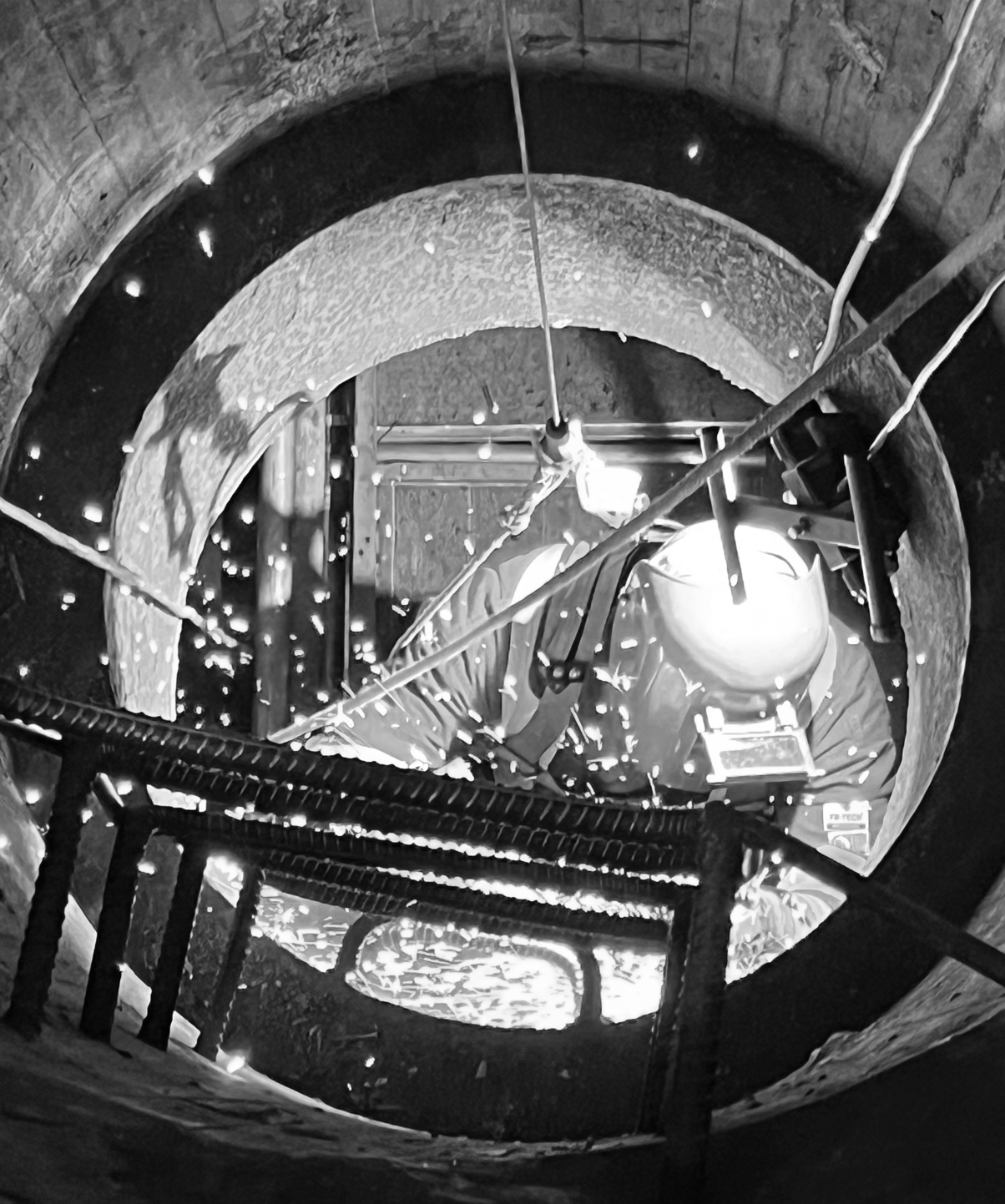 Worker inside a Confined Space performing welding wearing their protective gear inside a deep, circular well with scaffolding, pipes, and a light source illuminating the construction site. They are attached to fall restricting and arresting devices.