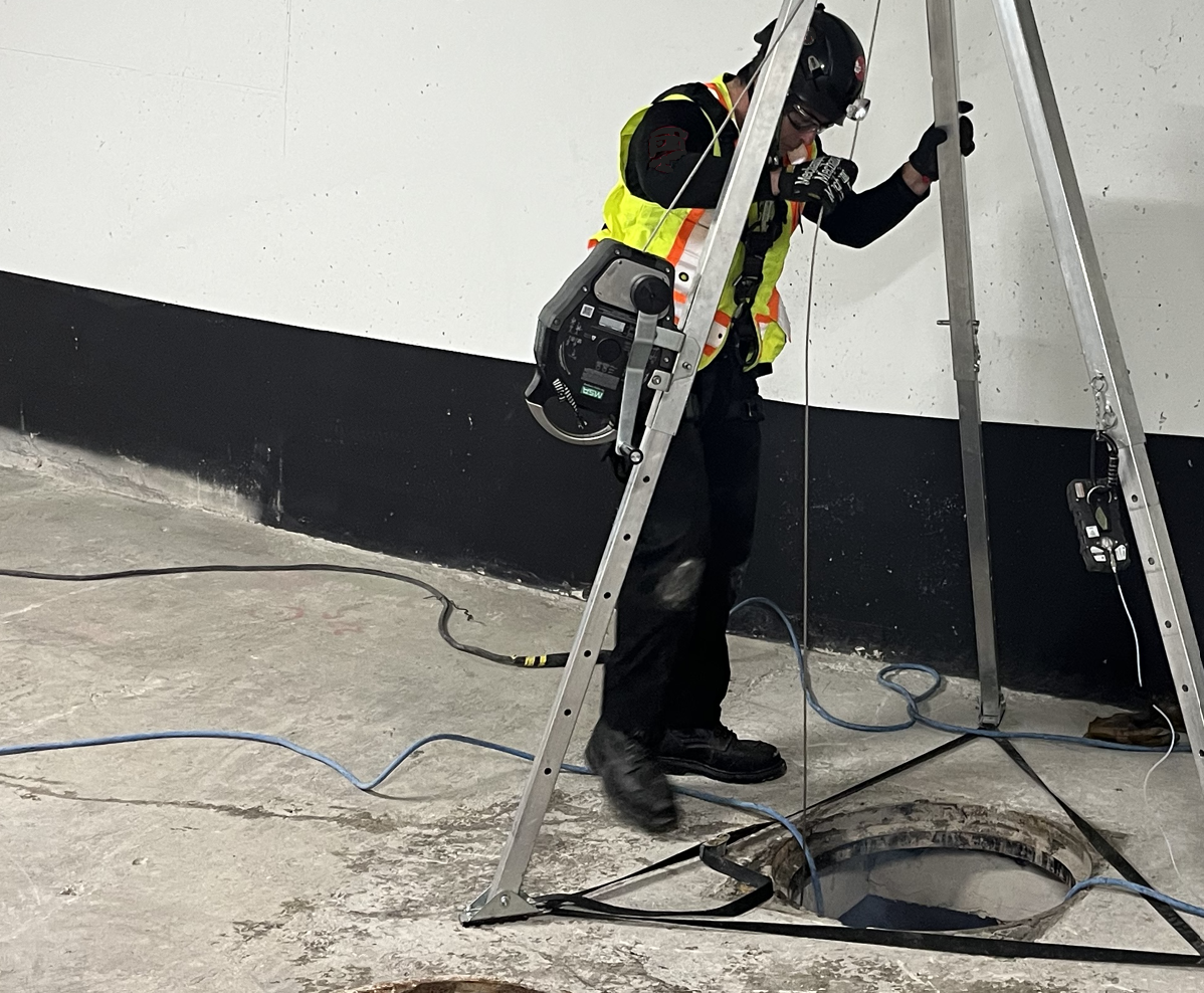 A worker wearing safety gear and a high-visibility vest inspects a large manhole opening with a ladder and equipment nearby.