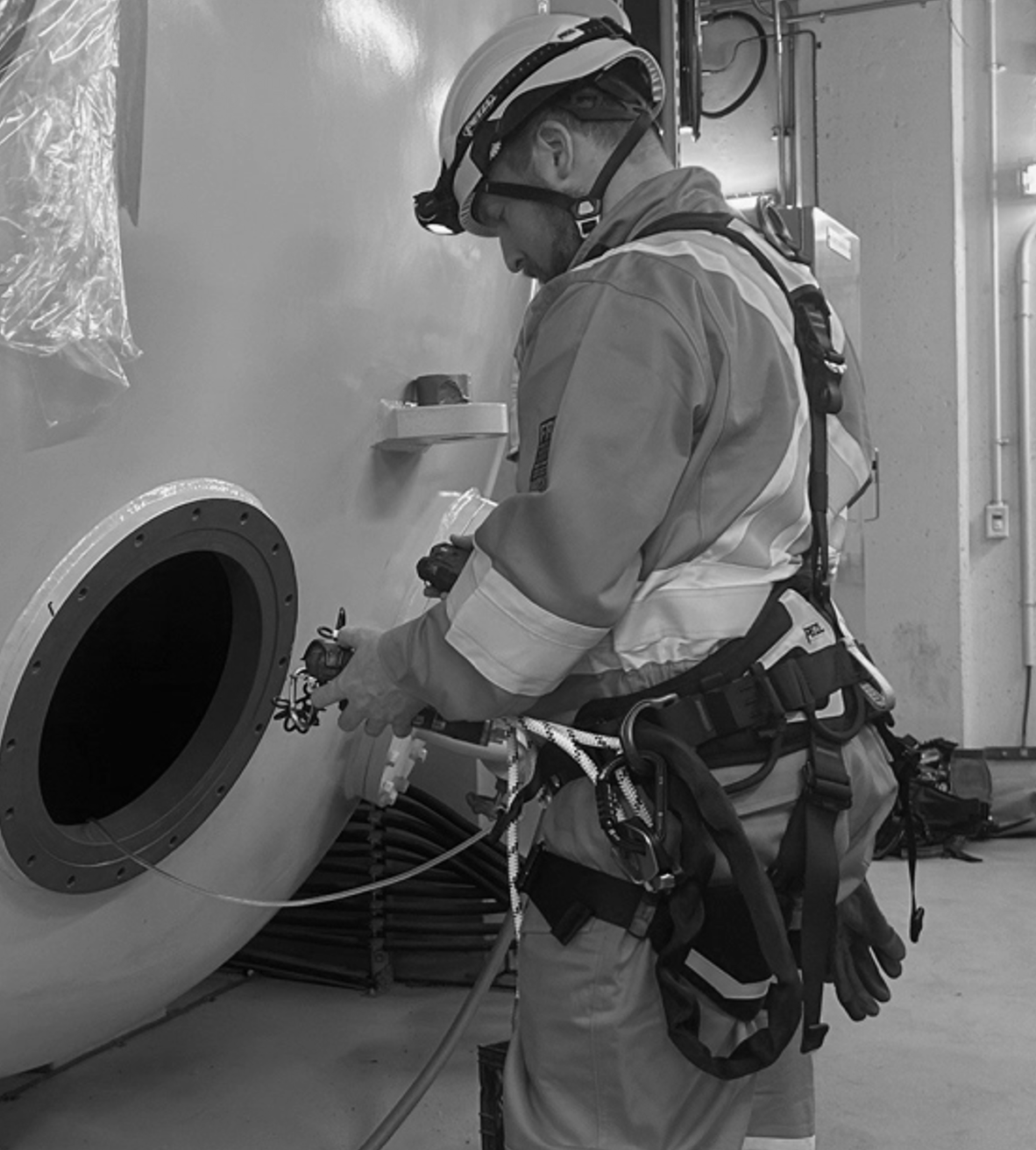 A worker in orange safety gear and yellow helmet kneeling on the floor, working near a large piece of industrial equipment. The worker is holding an air monitor sampling air within a confined space prior to a worker entering to ensure the air is safe