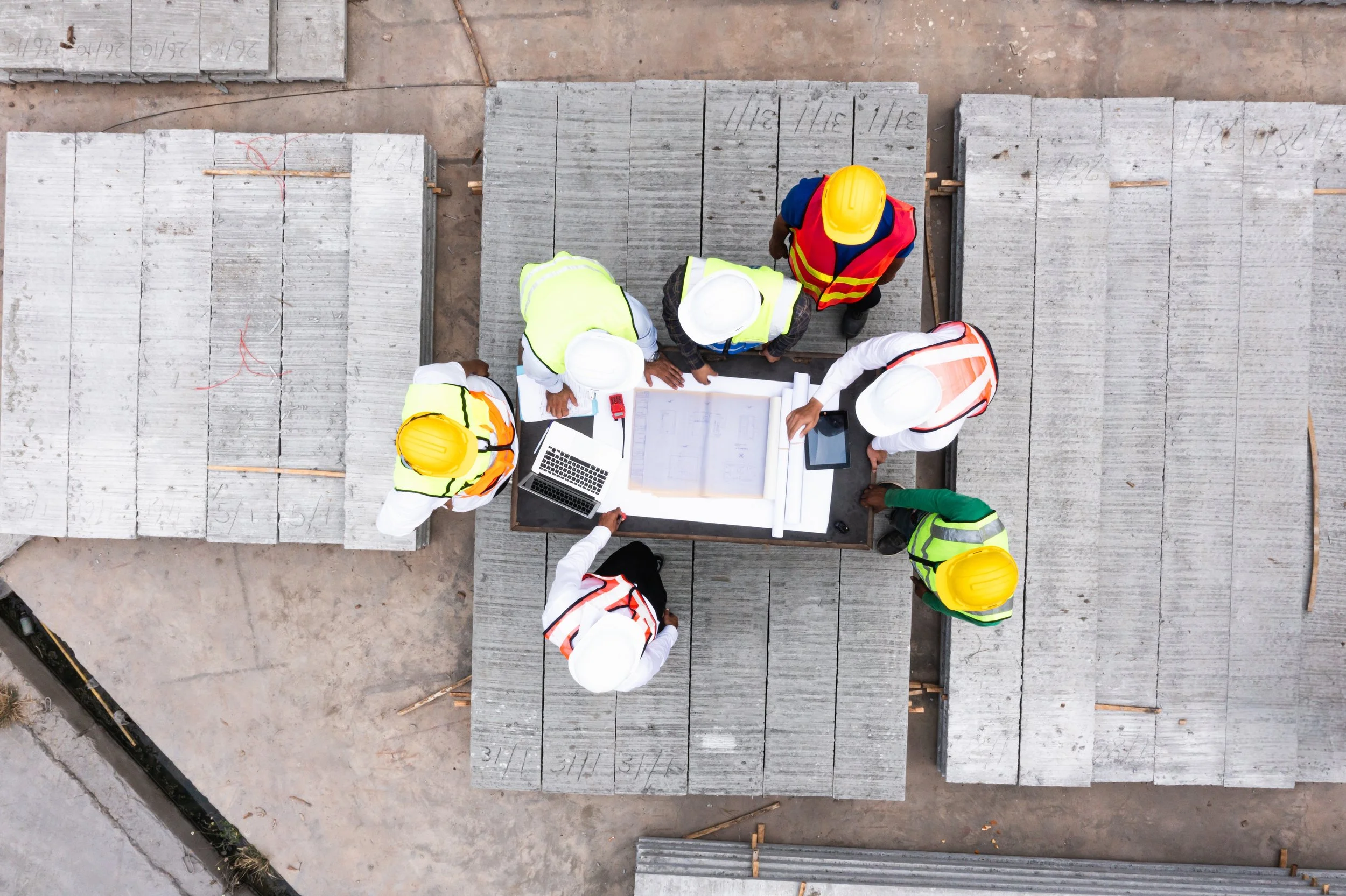 Top-down view of construction workers and health and safety consultants in safety vests and helmets gathered around a table on a construction site, reviewing documents and plans to creating solutions for health and safety hazards and risks.