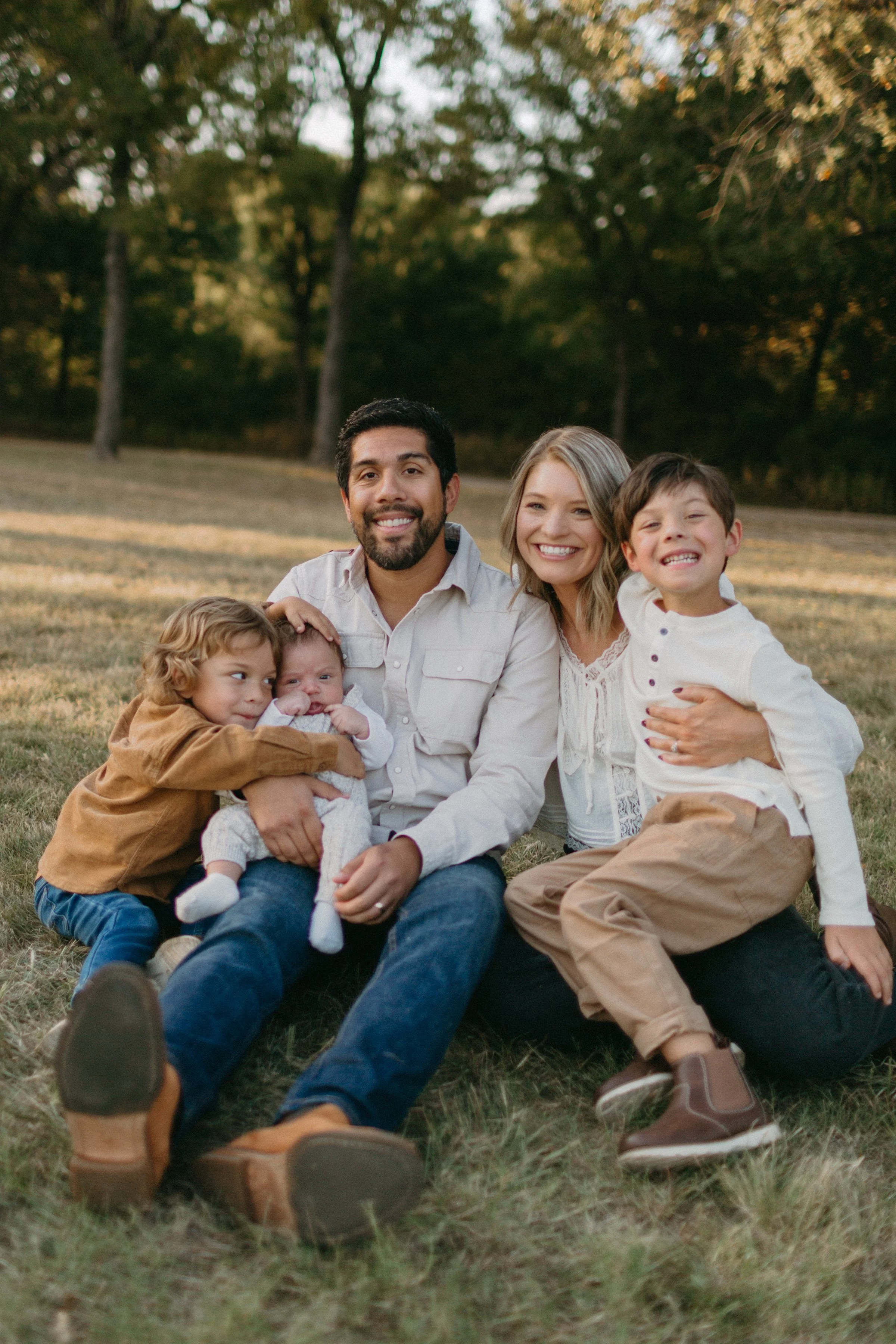Family of five sitting on the grass outdoors, smiling and enjoying each other's company in a park or natural setting during daytime.