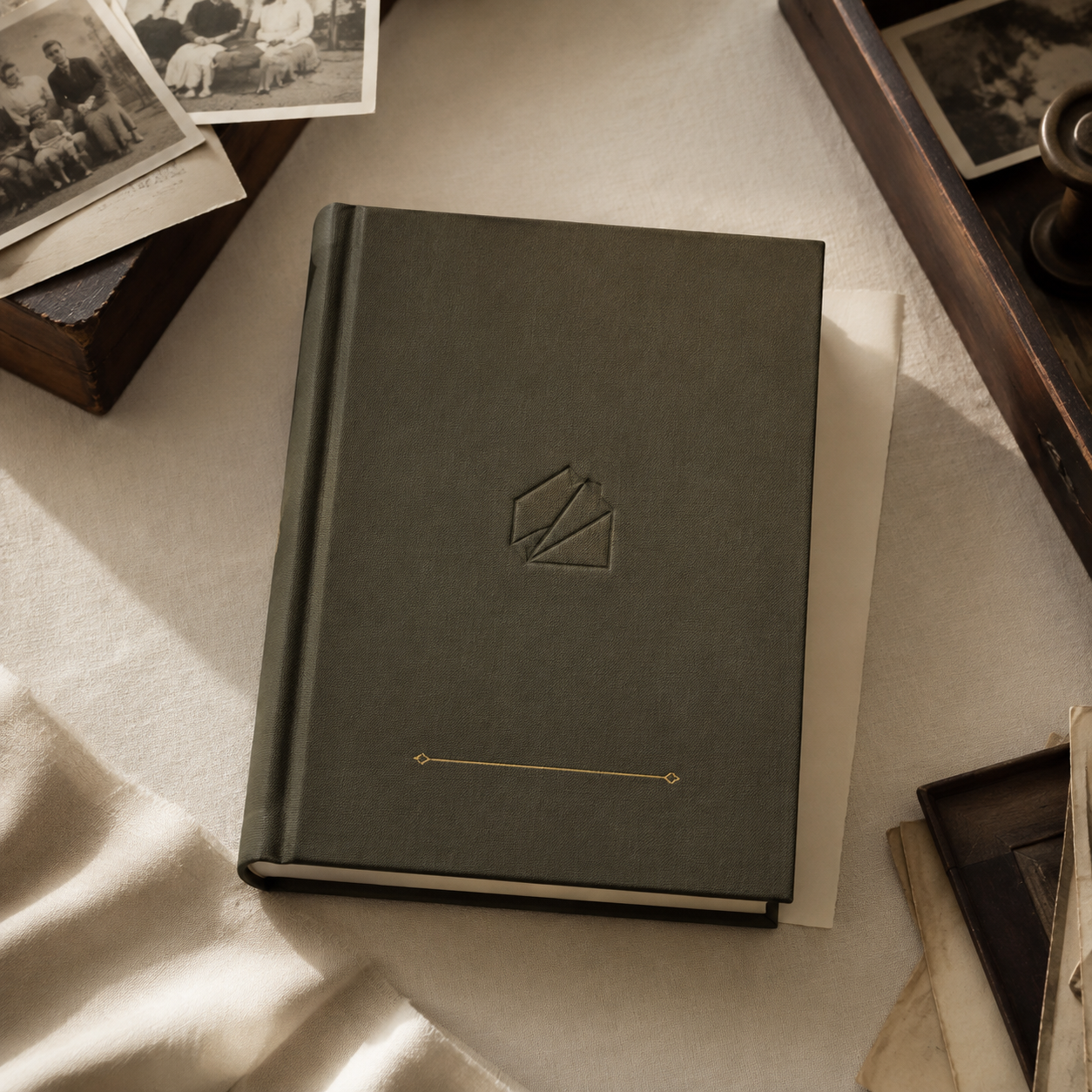A closed, dark green hardcover book with a geometric embossed logo on the cover, resting on a beige fabric surface. Surrounding the book are vintage black-and-white photographs and wooden trays with additional papers and objects.