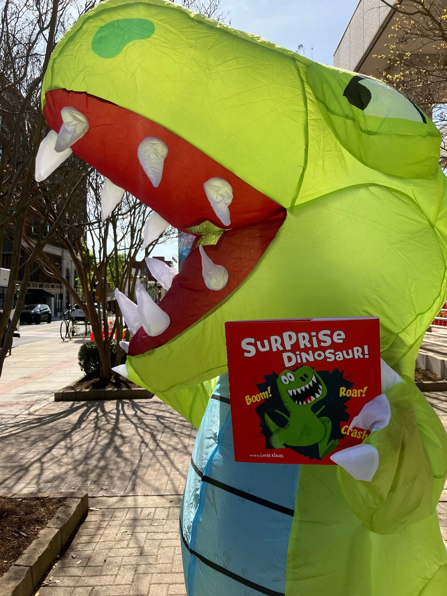 Colorful inflatable dinosaur costume with a bright green body, red mouth, and white teeth. The costumed person is holding a copy of the "Surprise Dinosaur' book.