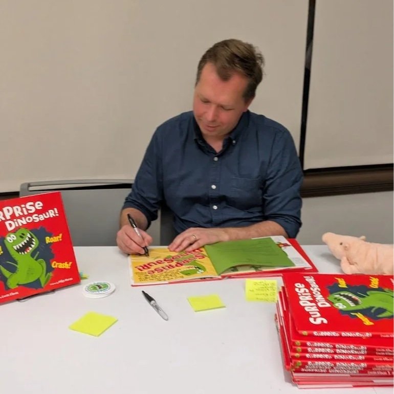 Author, Lucas Klauss, sitting at a table signing a copy of a children's book titled "Superprise Dinosaur!" with colorful illustrations of a green cartoon dinosaur on the cover. There are yellow sticky notes, a pen, and a stack of book on the table.