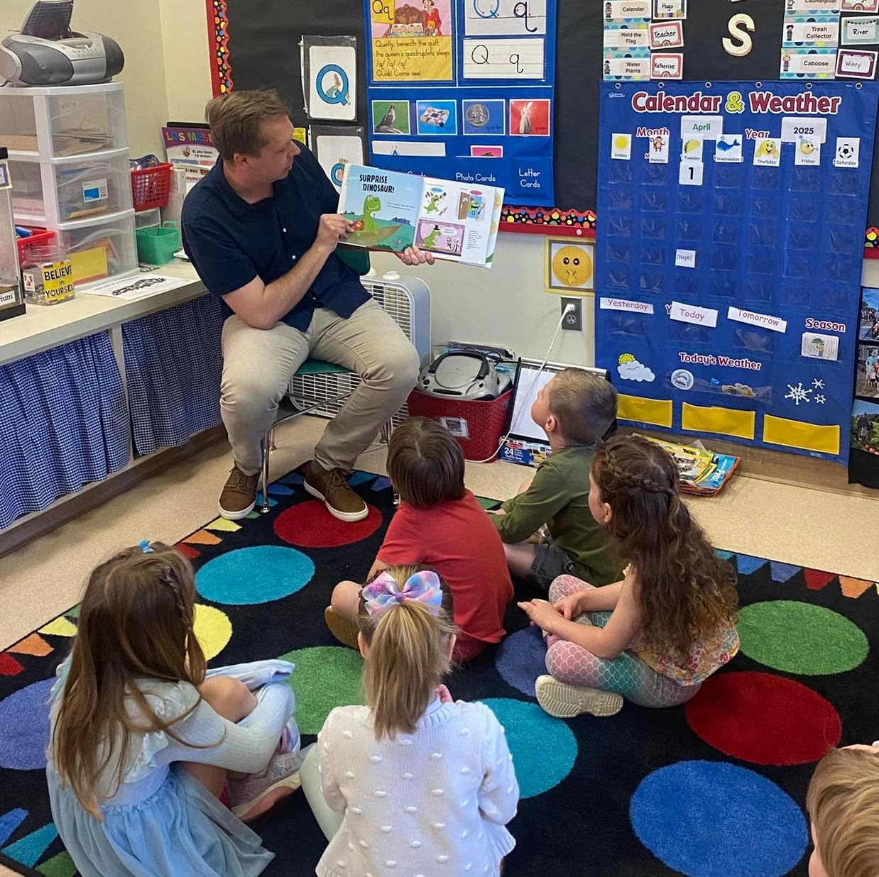 Author, Lucas Klauss, reading a children's book titled 'Surprise Dinosaur!' to a group of young students seated on a colorful polka-dot rug in a classroom. The classroom has educational posters and a calendar on the wall.