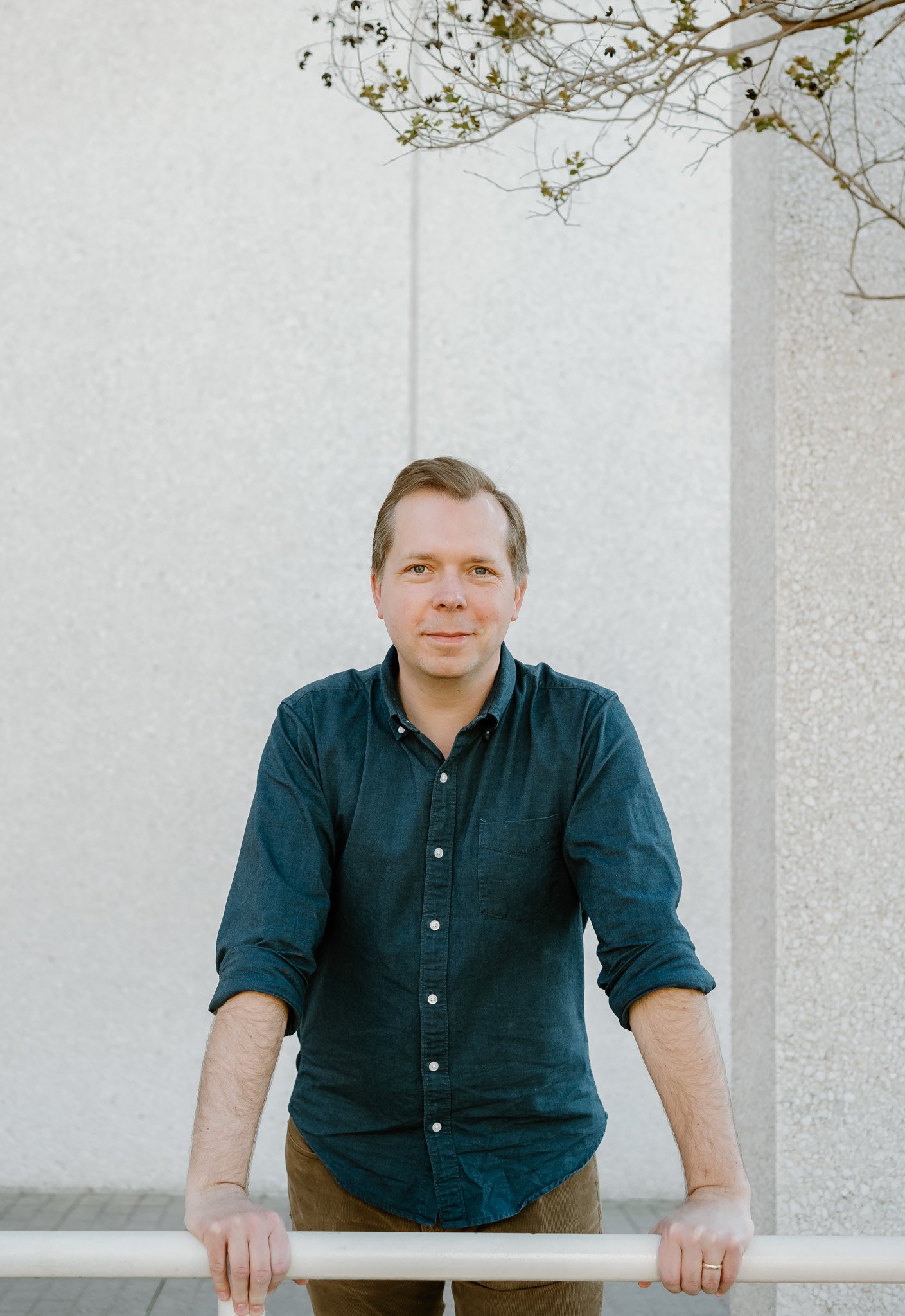 Author, Lucas Klauss, wearing a navy blue button-up shirt and brown pants is standing outdoors, leaning forward with his hands on a white railing. Behind him is a textured light-colored wall and a few branches of a small, leafless tree.