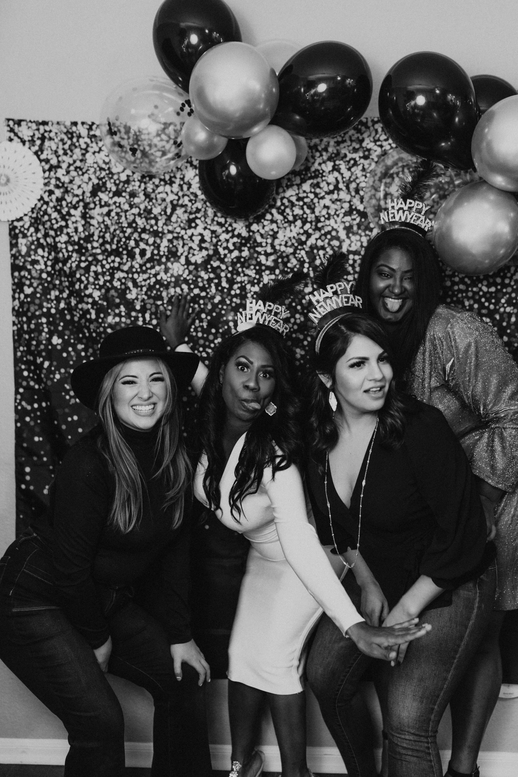 Group of women celebrating New Year, wearing 'Happy New Year' party hats and making fun faces, with balloons and confetti background.