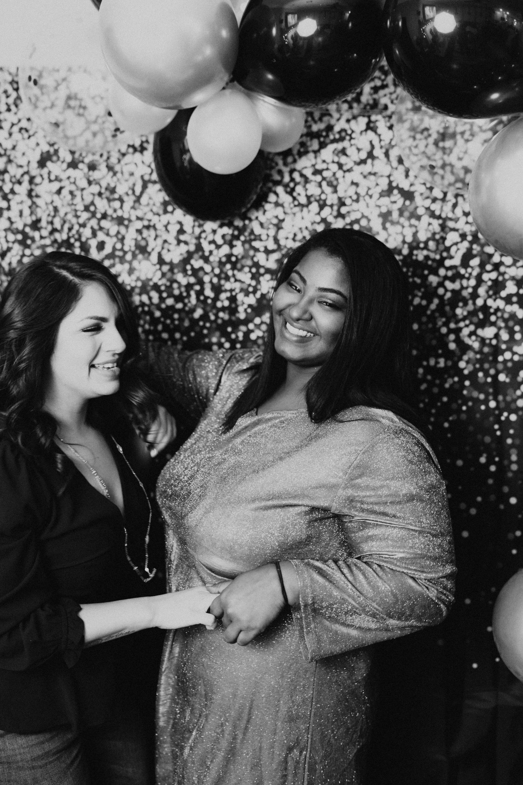Two women celebrating at a party with balloons and confetti in the background, one wearing a shiny dress and the other wearing a dark top.