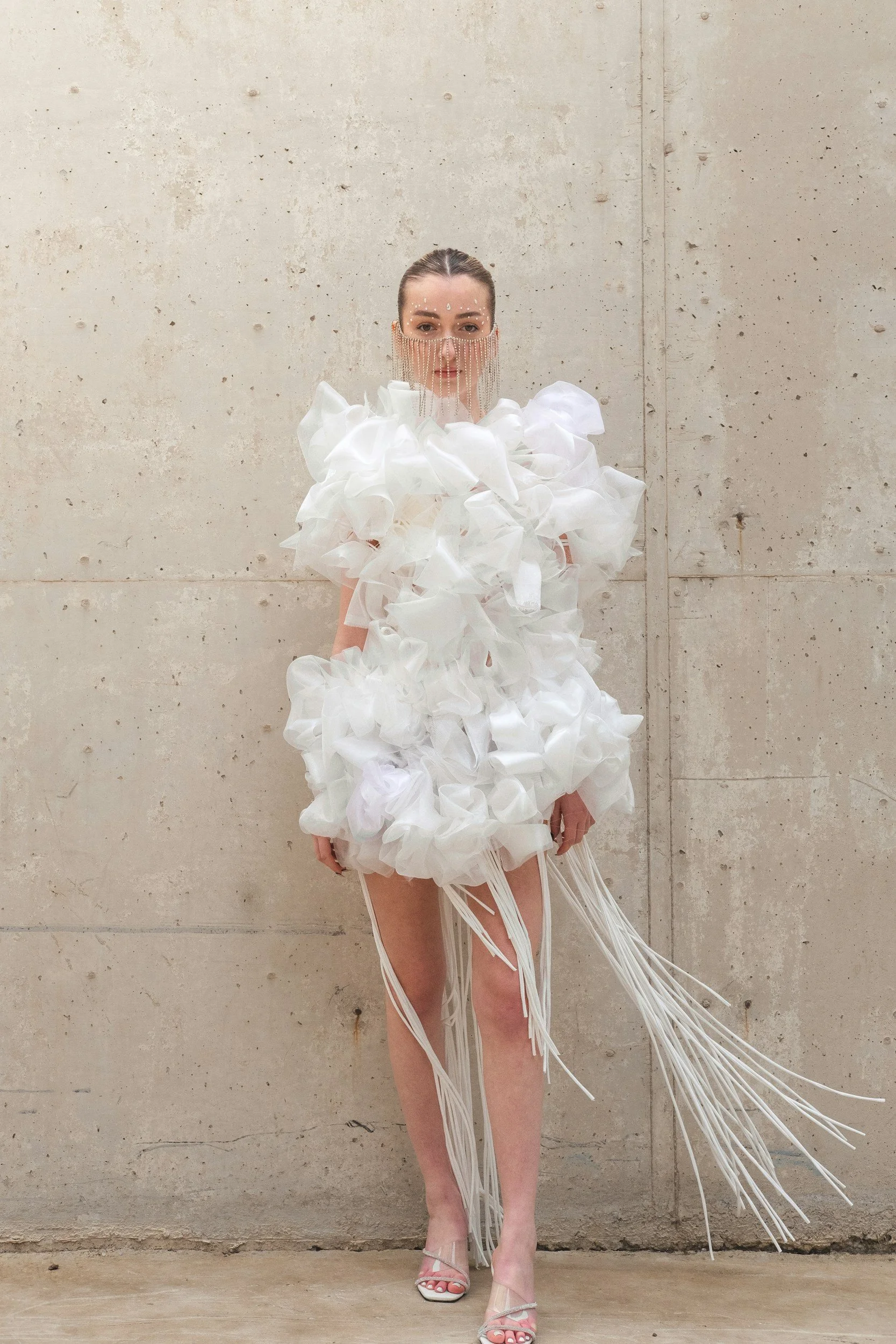 A woman wearing a white avant-garde dress made of large, fluffy fabric with long white ribbons hanging down, standing against a plain concrete wall.