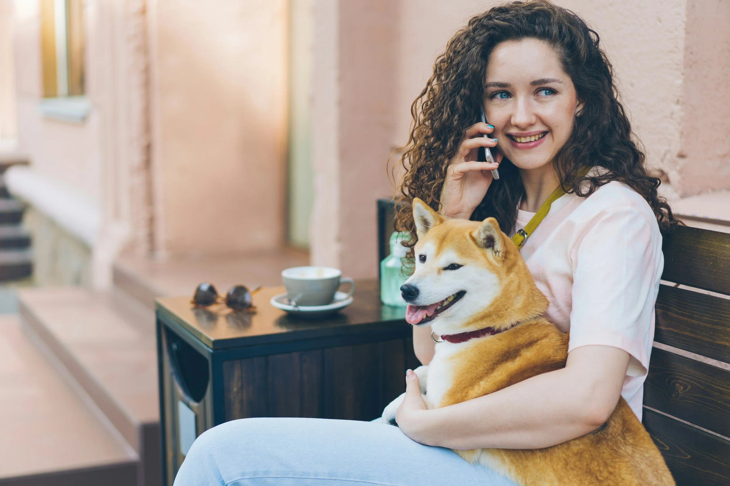 A young woman with curly brown hair and blue eyes smiling while talking on her cellphone, sitting on a bench with a Shiba Inu dog in her lap at a cafe or outdoor seating area.