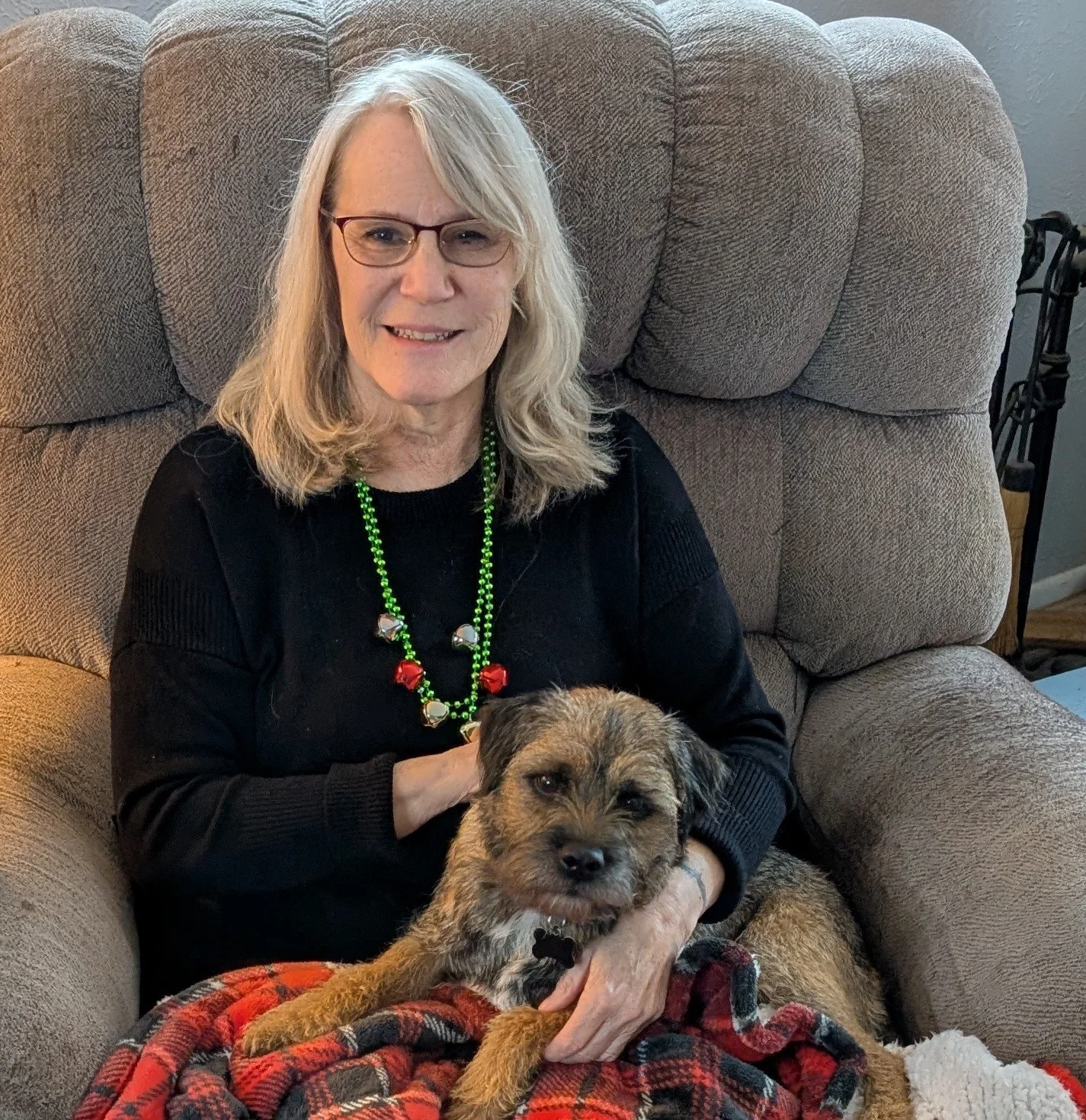 A woman with blonde hair and glasses sitting on a beige recliner, holding a small, brown, black, and gray dog with a red and black plaid blanket in her lap.