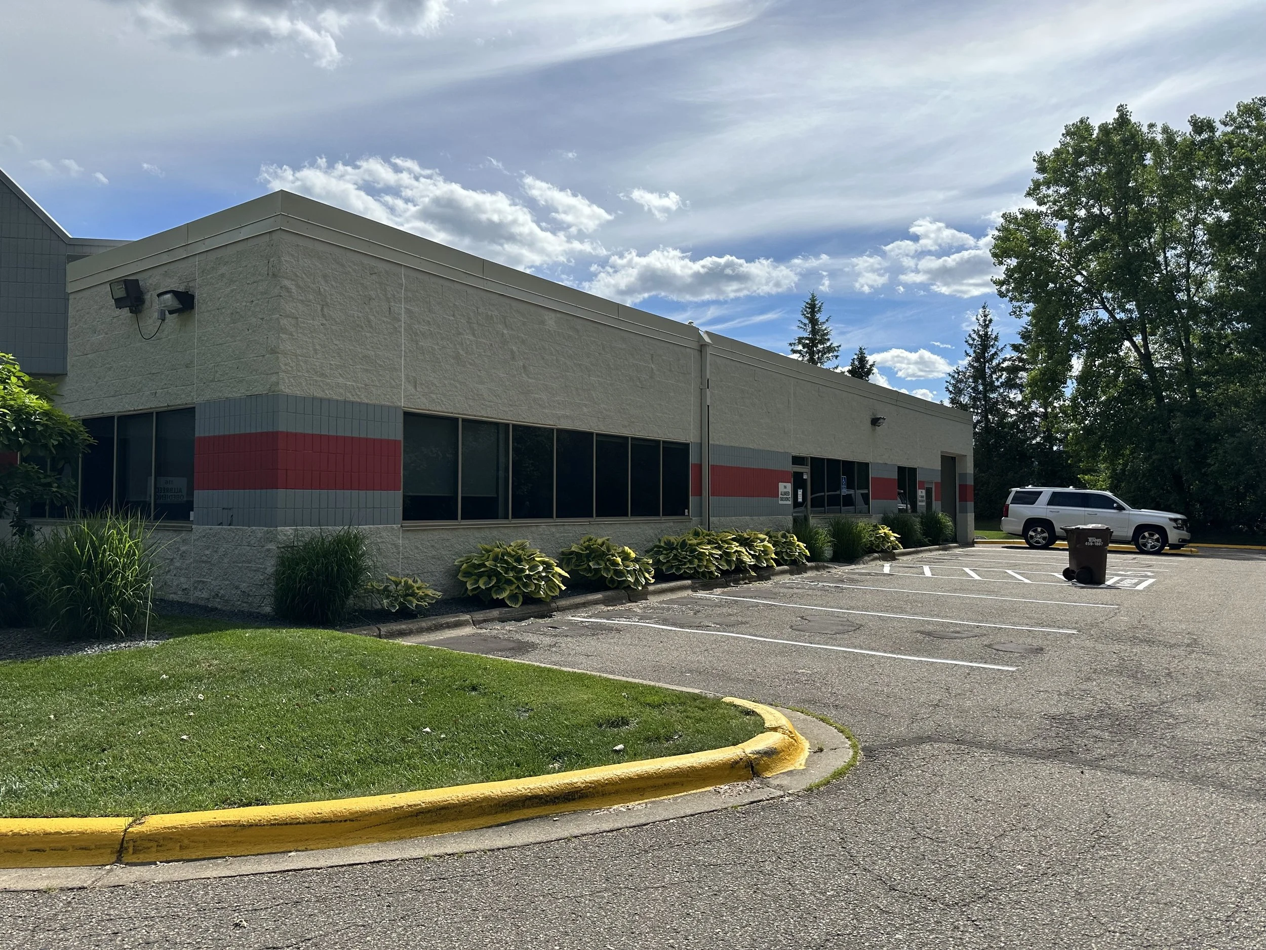 A commercial building with gray exterior walls, large windows, and a red stripe running along the lower part. There are parking spaces with a white vehicle and a trash bin in the parking lot. The background shows tall trees and a partly cloudy sky.