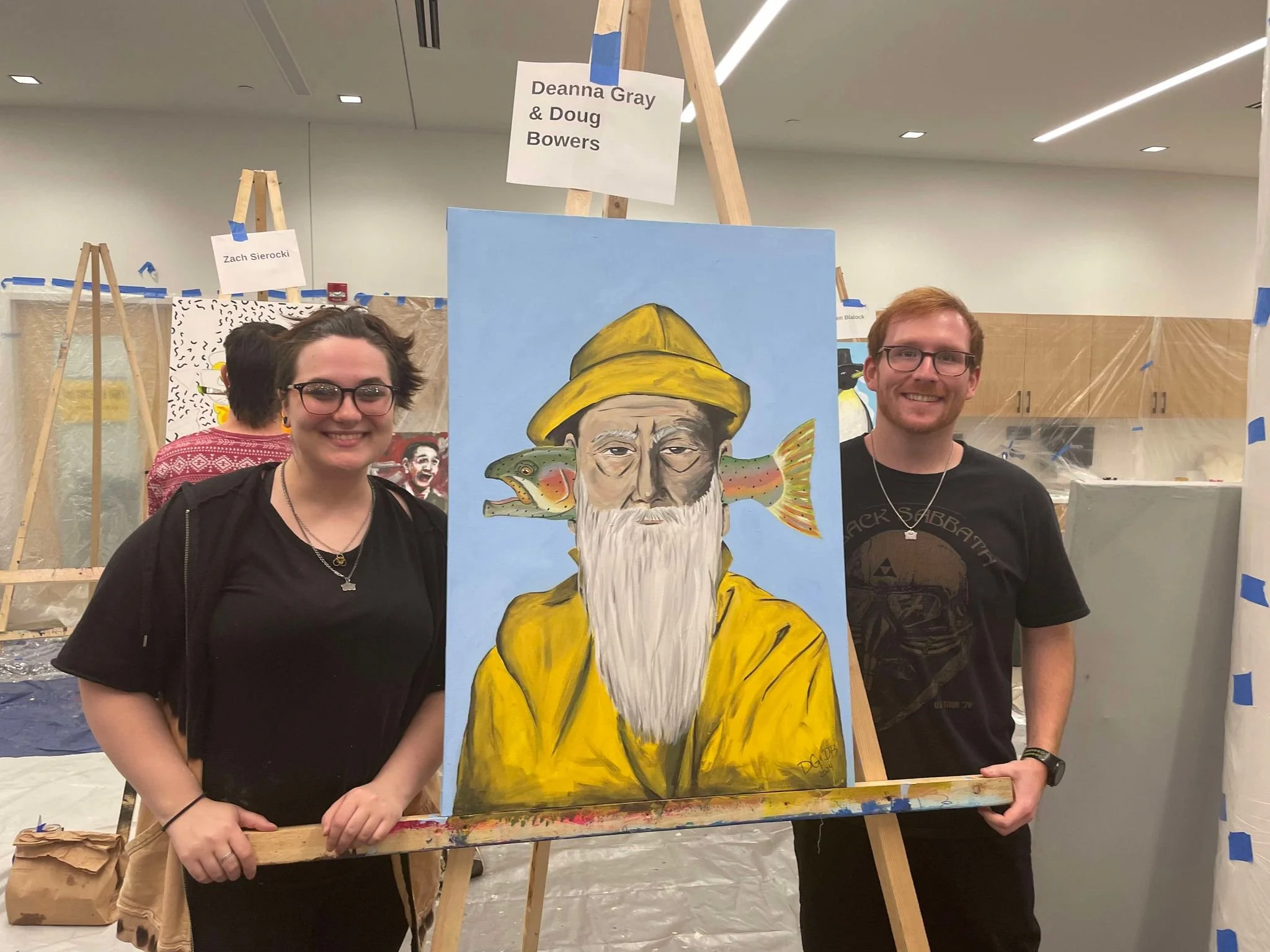 Two people standing on either side of a colorful painting of an elderly man with a beard, a yellow coat, and a yellow hat, with digital art elements of a frog and fish around the man. They are in an art gallery or studio setting, with art easels and 