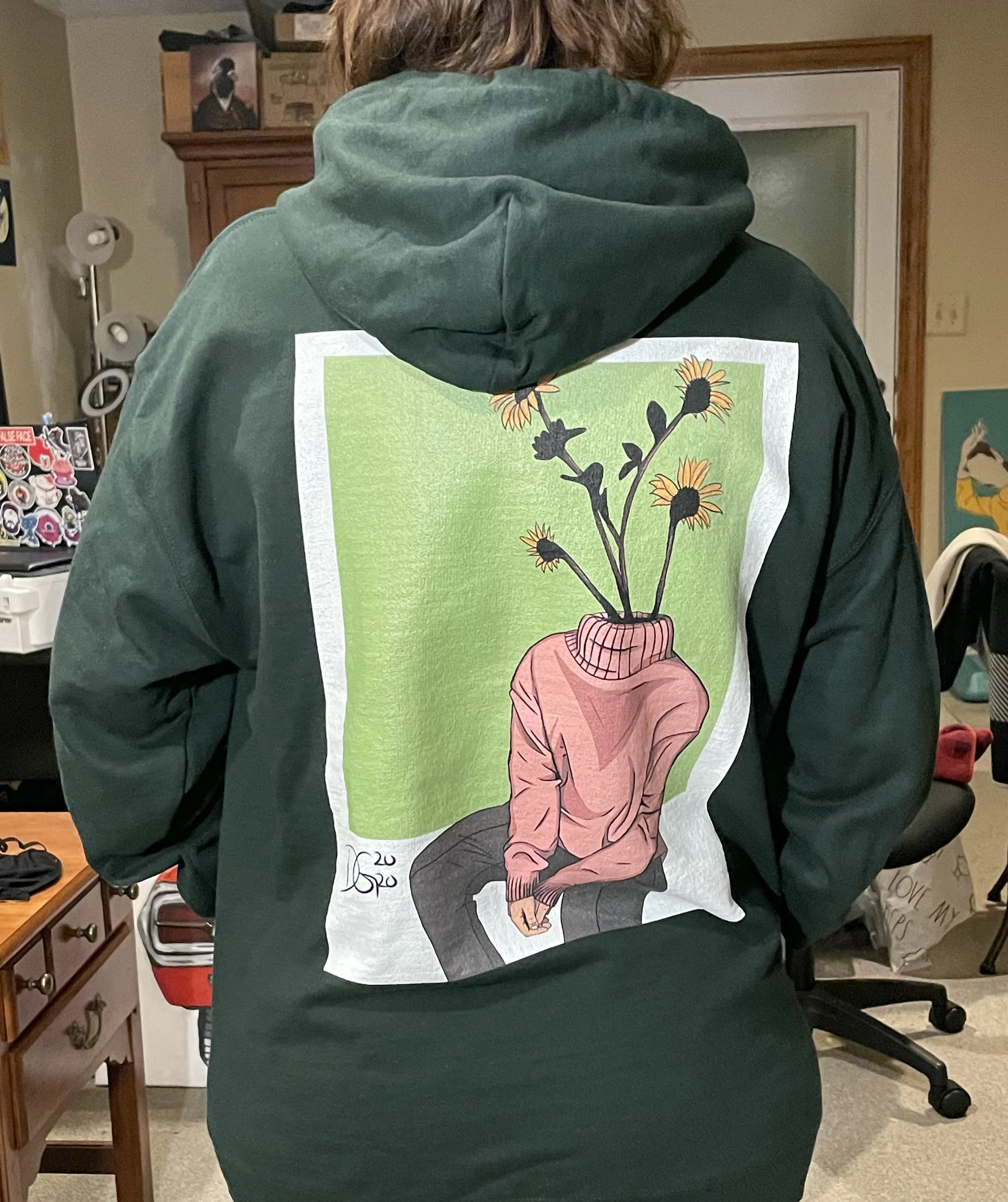 Back of a person wearing a hoodie featuring a colorful illustration of a person with a sunflower in a pot on their head. The background contains a kitchen area with a white door, a wooden cabinet, and a small collection of stickers on a surface.