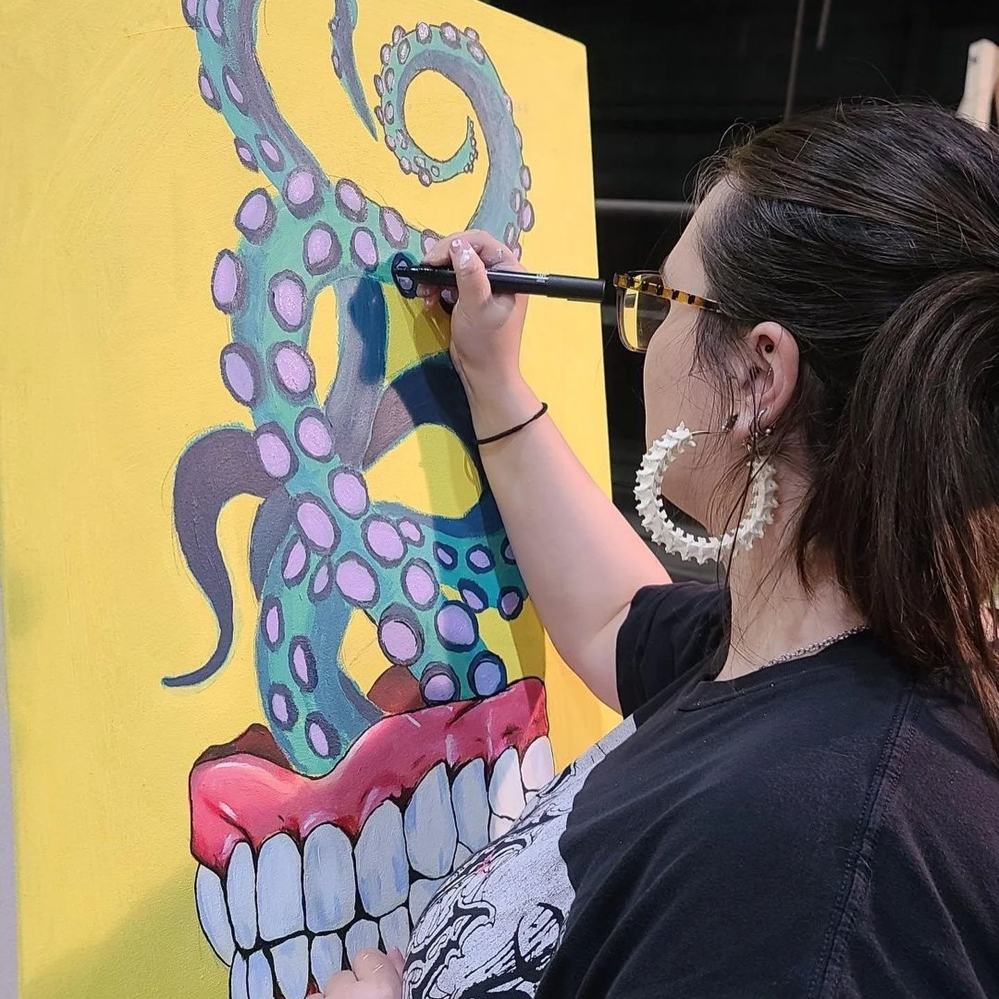 A woman with dark hair and glasses is painting a colorful skull on a yellow background. The skull features a turquoise octopus with purple spots coming out of the top of the head.