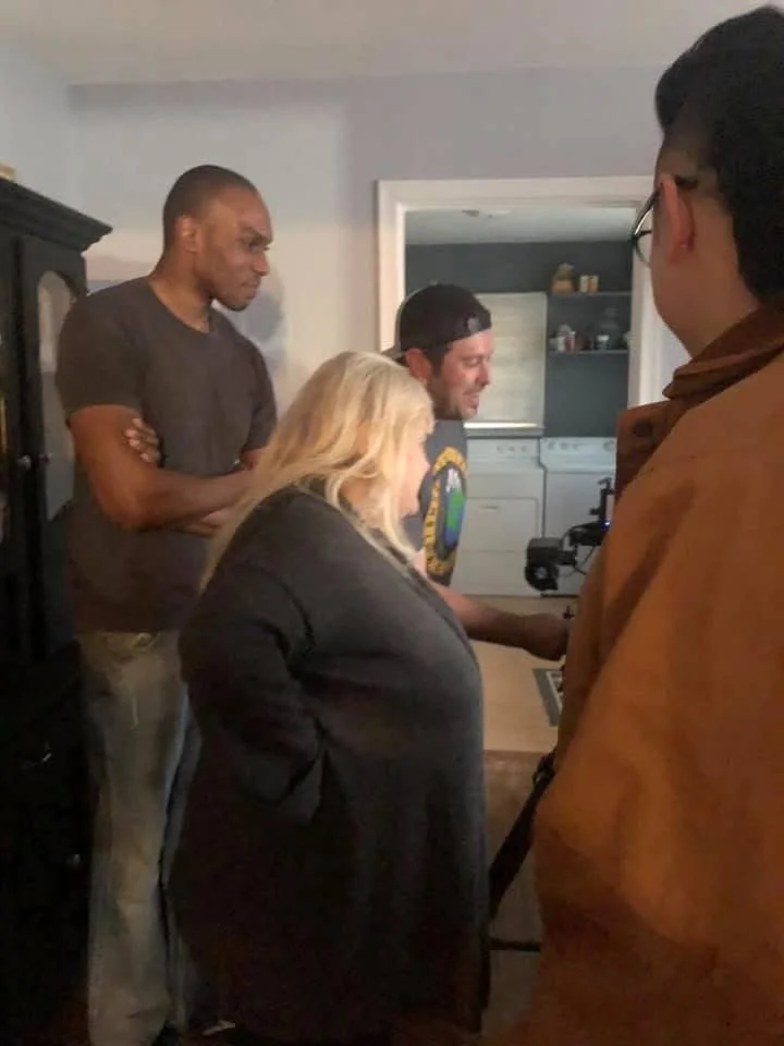 Four people, three men and one woman, are gathered around a table looking at a laptop in a home setting.