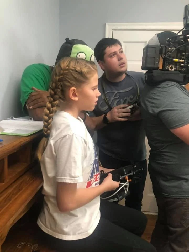 A girl with braided hair holding a remote control in a room with two young men, one with a gaming controller and the other behind a professional camera, watching a filming scene.