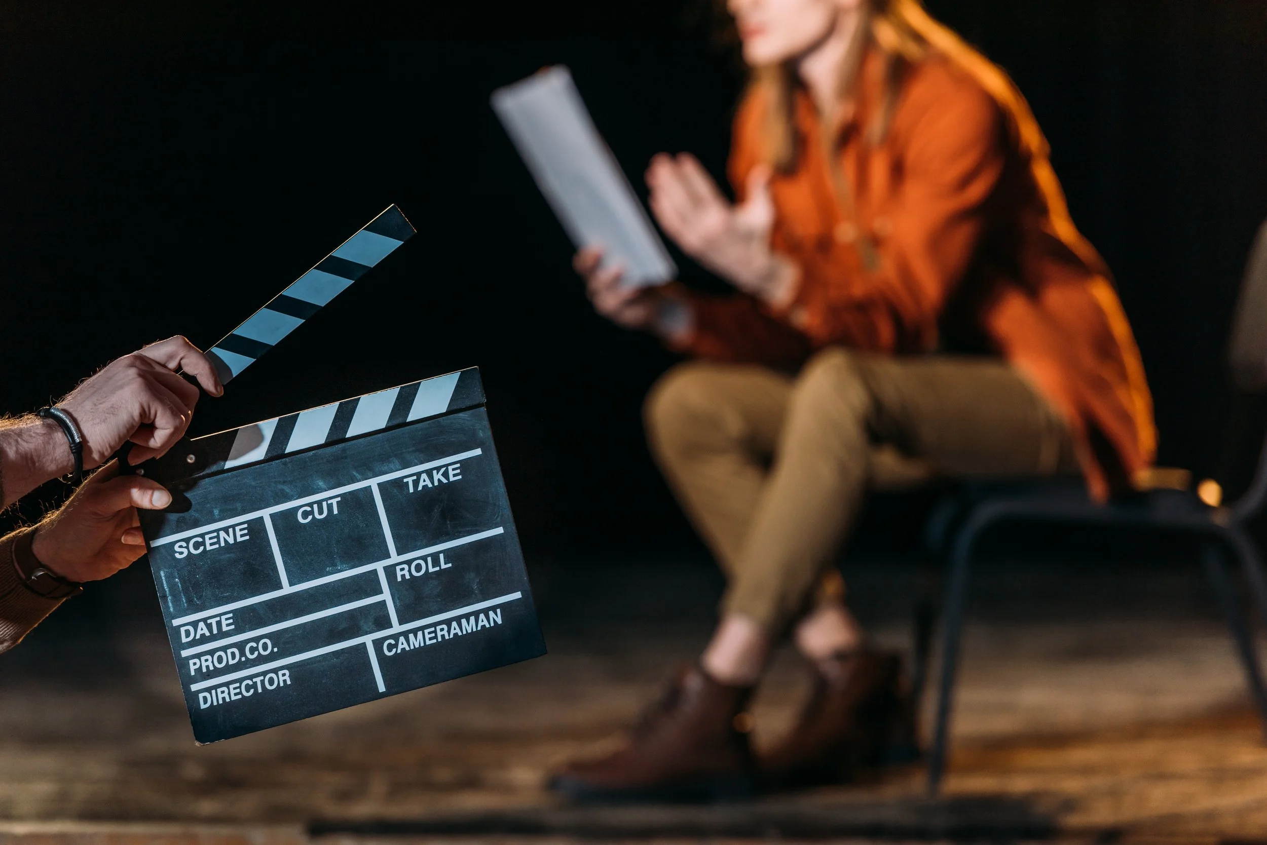 A film director's clapboard held in front of a woman in an orange shirt, with a blurred background of a person sitting on a chair, wearing a red shirt, in a dark room.
