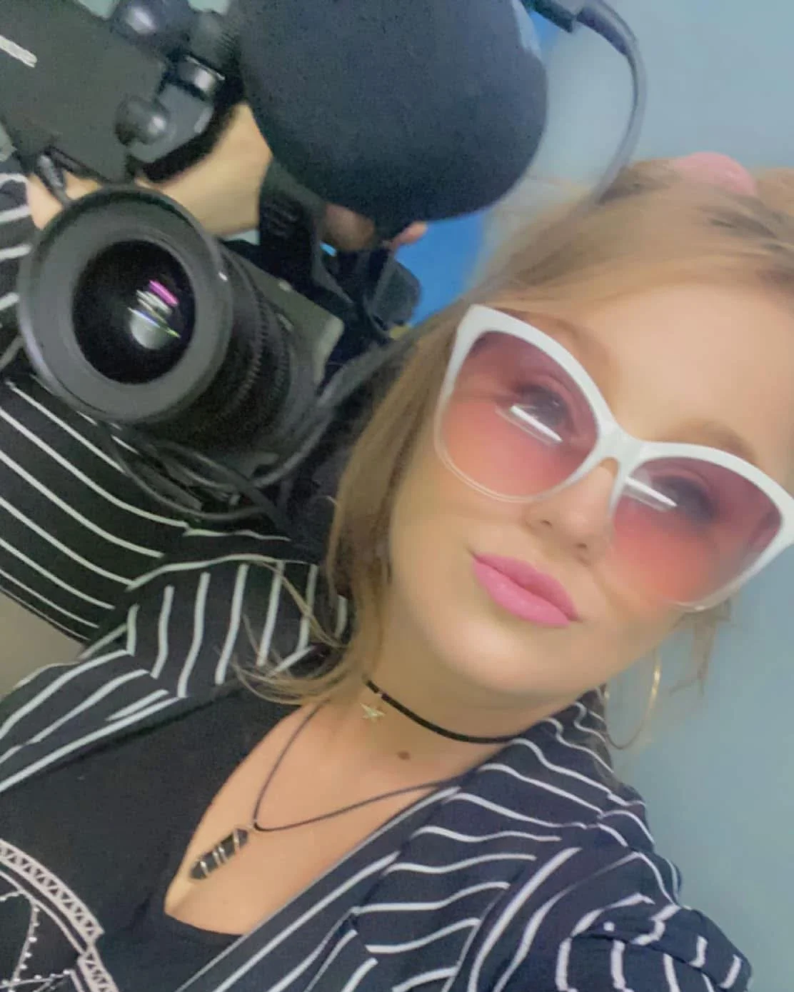 Close-up of a woman wearing large pink sunglasses, a black choker, necklaces, and a striped jacket, holding a professional camera with a large lens.