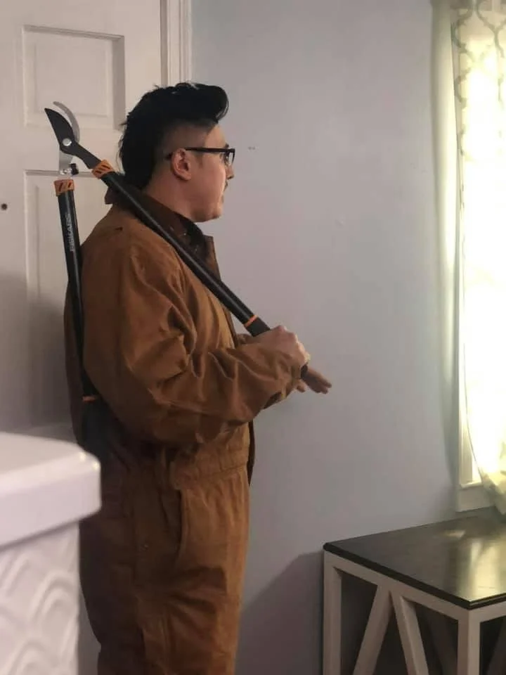 A person wearing glasses and a brown jumpsuit, holding an axe over their shoulder, standing indoors near a window with yellow curtains.