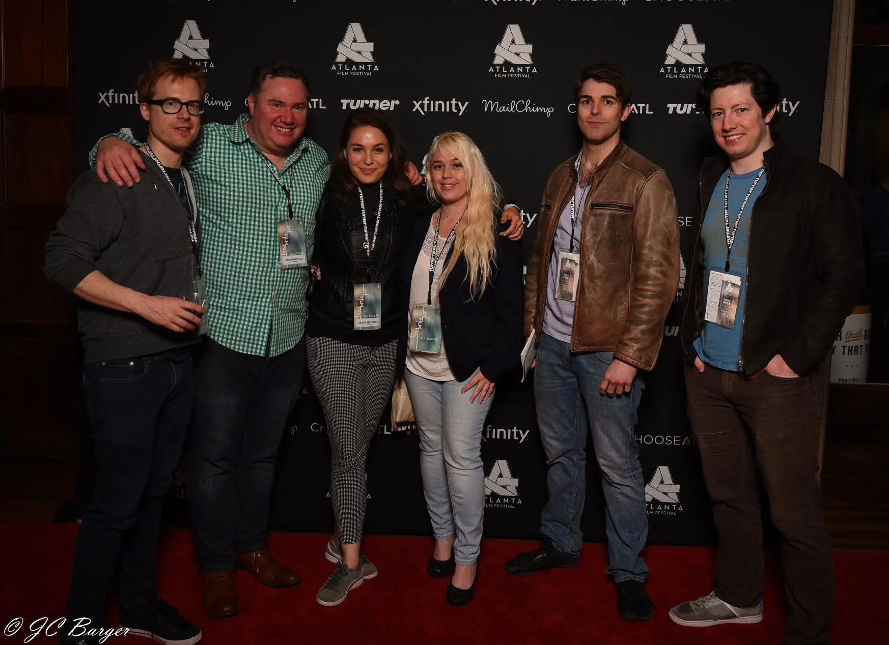 Six people posing together in front of a step and repeat banner at the Atlanta Film Festival. They are smiling and wearing event badges around their necks. Four men and two women, dressed in casual to semi-formal attire.