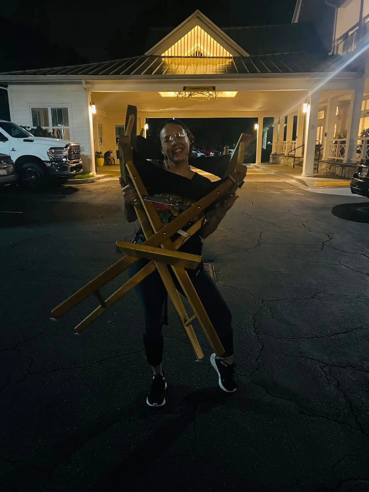 A person holding a wooden chair outdoors at night, standing on a parking lot with cars, near a building with illuminated interior lighting.
